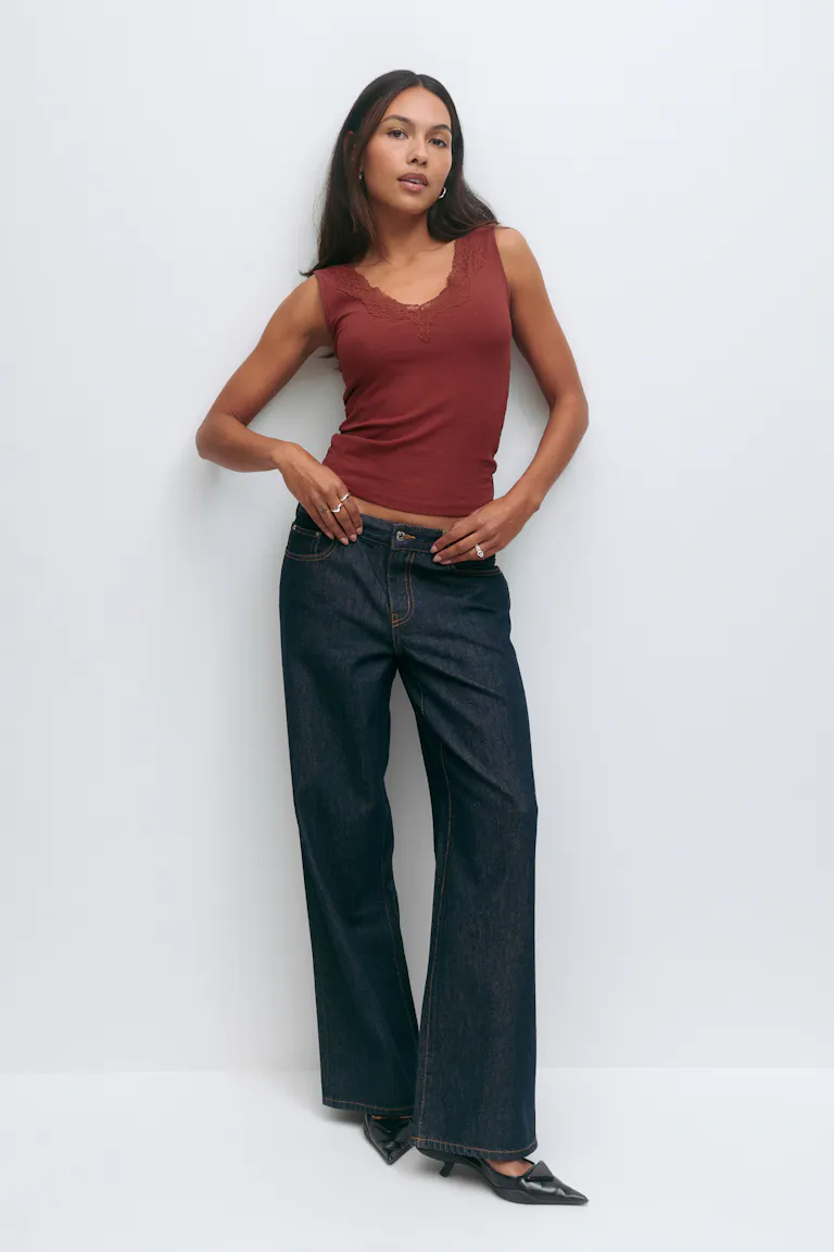 Mouwloze, roestkleurige tank met V-hals met kanten rand, donker indigo jeans met hoge taille en zwarte hakken met spitse teen.