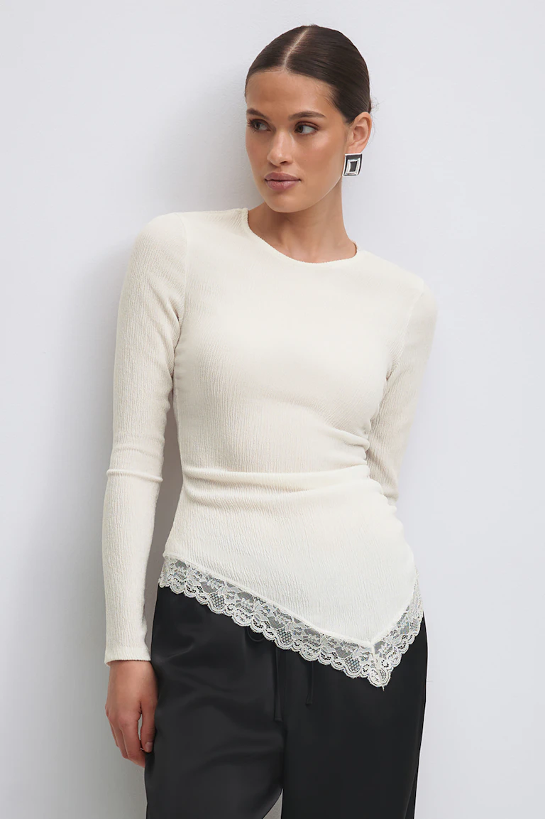 Een crèmekleurige geribde, getailleerde top met lange mouwen en een puntige, met kant afgezette asymmetrische zoom, gecombineerd met een broek van zwart satijn met tunnelkoord en vierkante, zilveren oorbellen.