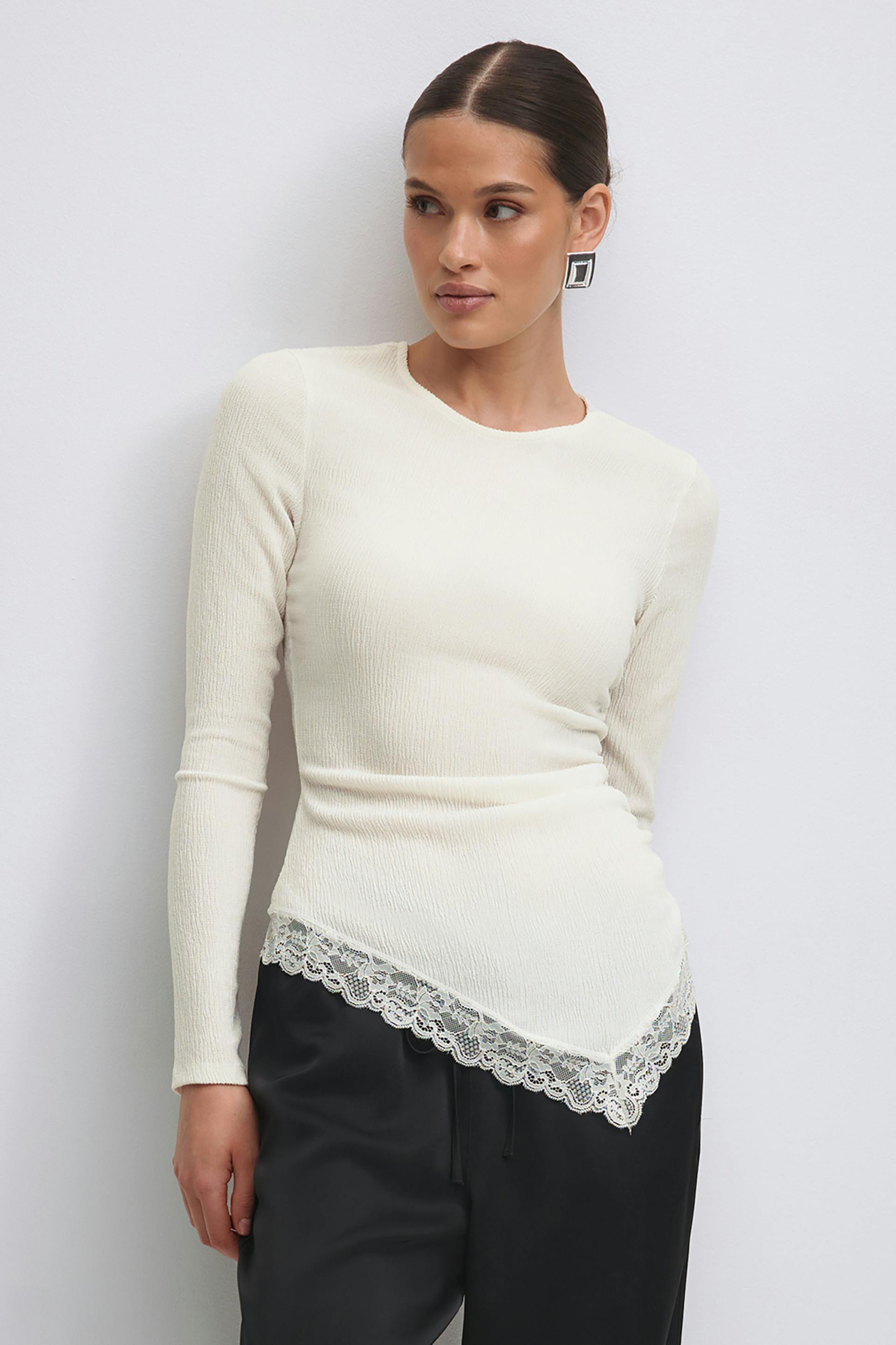 Cream ribbed long-sleeve fitted top with a pointed, lace‑trimmed asymmetric hem, paired with black satin drawstring trousers and square silver statement earrings.