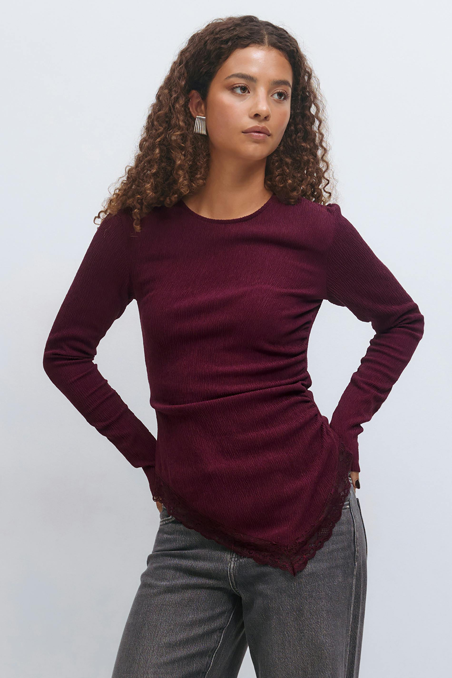 Long-sleeve textured burgundy top with a crew neckline and asymmetrical lace‑trimmed pointed hem; faded grey high‑waist jeans.