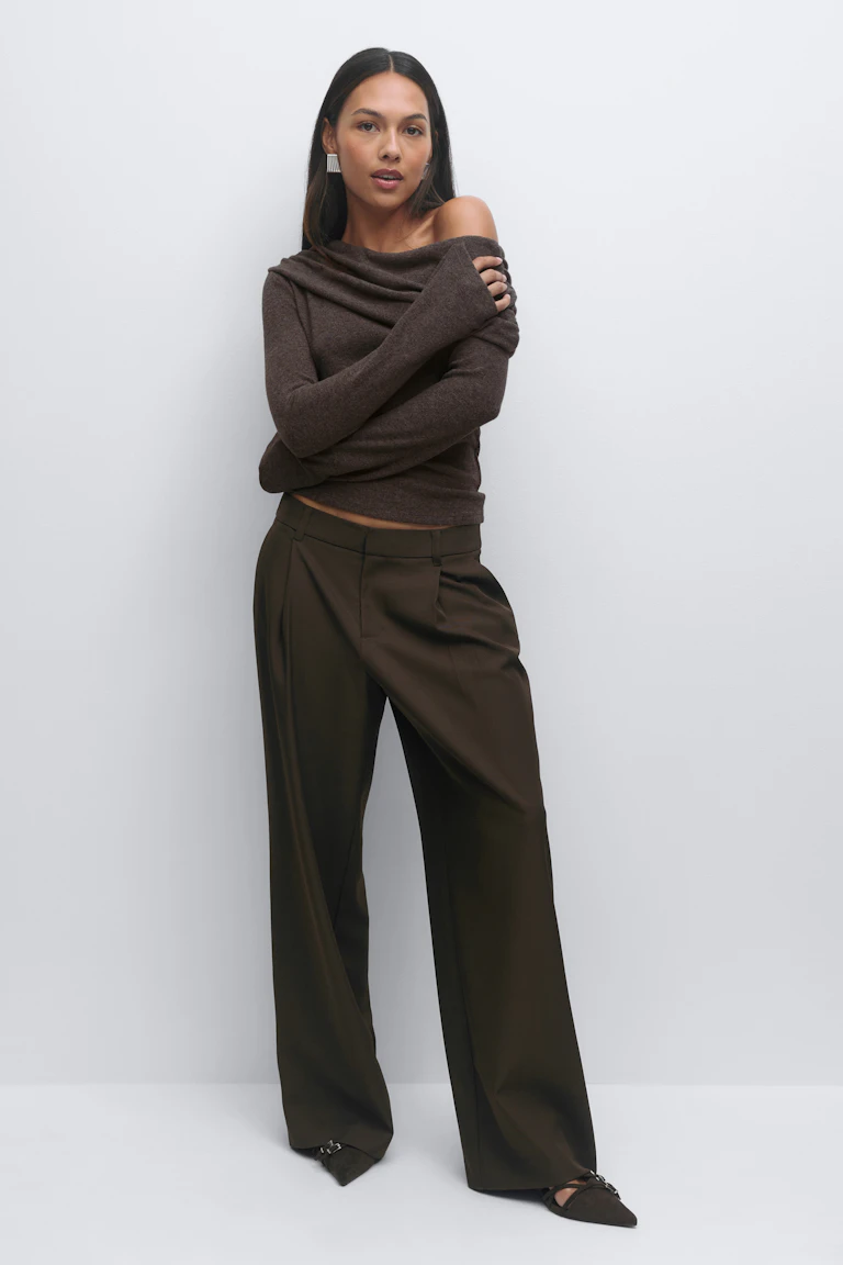 Donkerbruine gedrapeerde off-the-shoulder trui met lange mouwen, bijpassende wijde broek met hoge taille en plooien aan de voorkant, spitse platte schoenen met gespdetail en zilveren oorbellen.