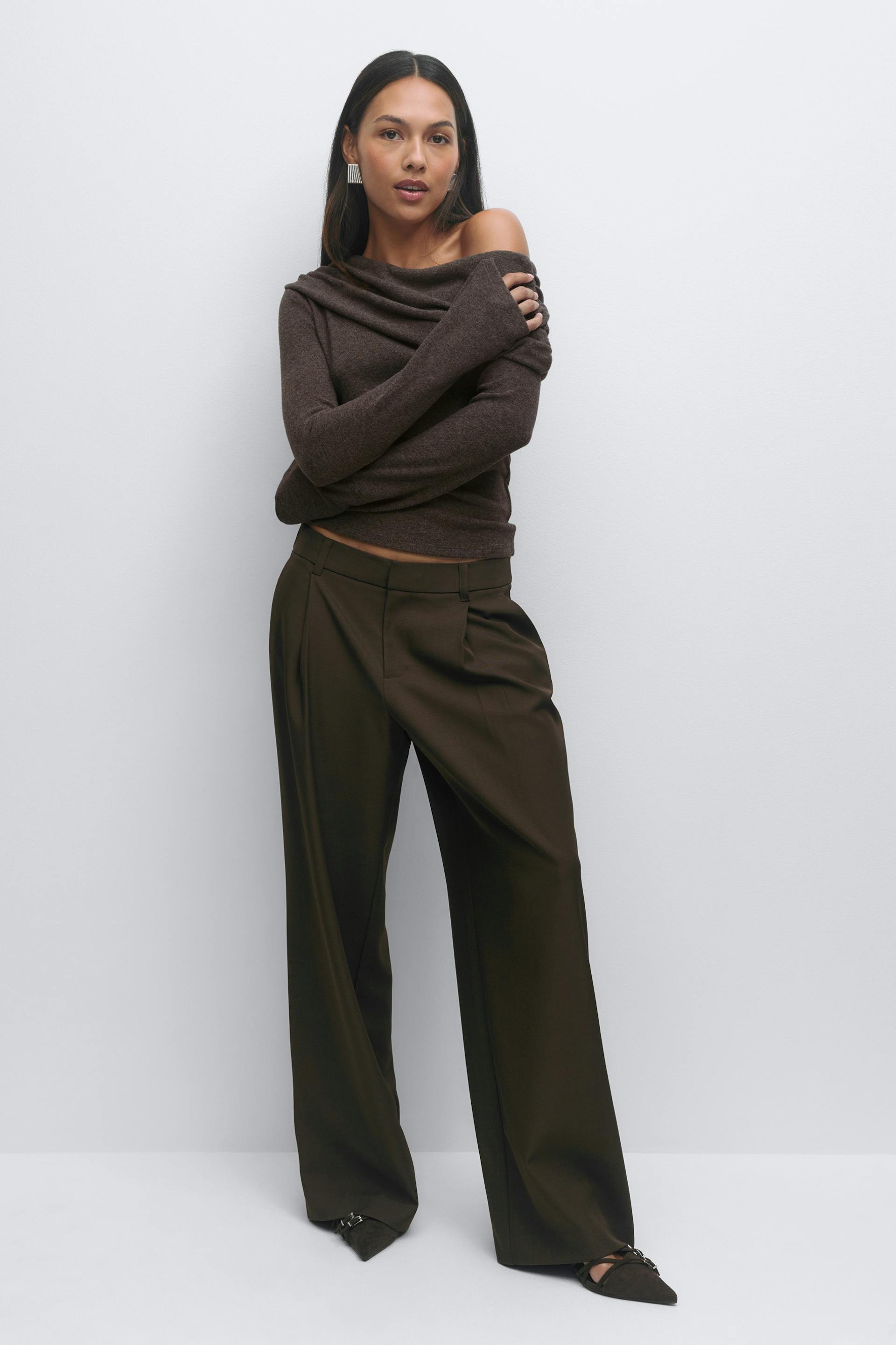 Dark brown off-the-shoulder draped long-sleeve sweater, matching high-waisted wide-leg trousers with front pleats, pointed-toe flats with buckle detail, and silver statement earrings.