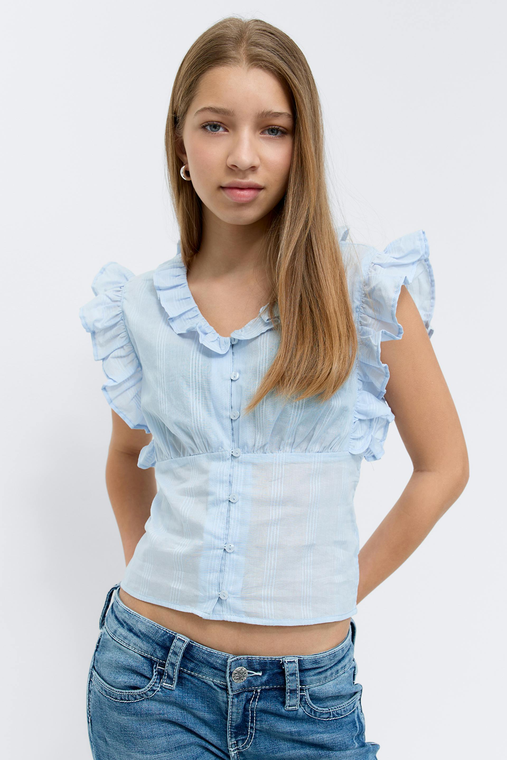 Light blue cropped blouse with ruffled collar and flutter sleeves, subtle vertical stripe texture and front buttons, paired with mid‑blue denim jeans and small silver hoop earrings.