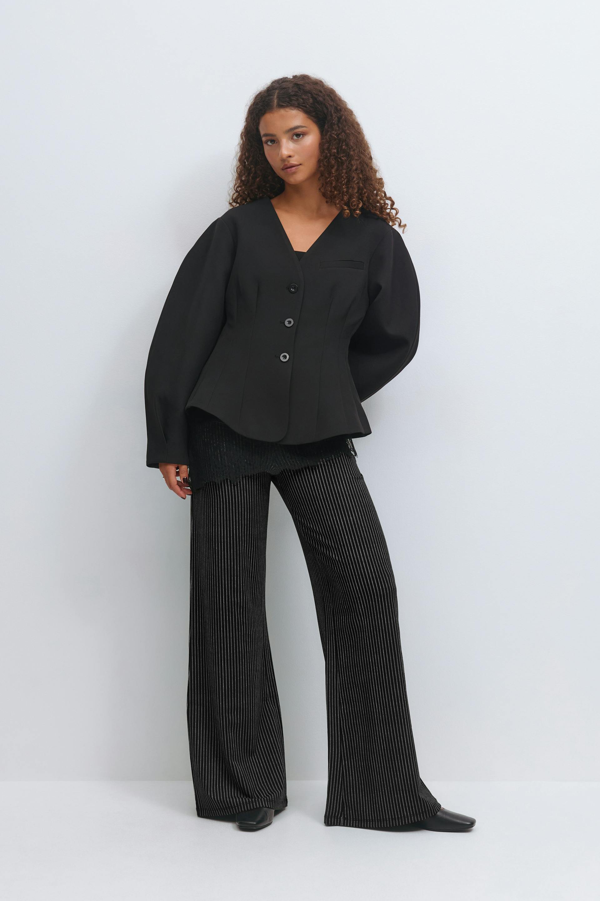 Musta räätälöity V-kauluksinen bleiseri, jossa on kolme nappia, istuva vyötärö ja tuuheat pitkät hihat, kerrostettu mustan pitsikoristeisen topin päälle; yhdistettynä mustiin nastaraidallisiin leveälahkeisiin housuihin ja mustiin teräväkärkisiin kenkiin.