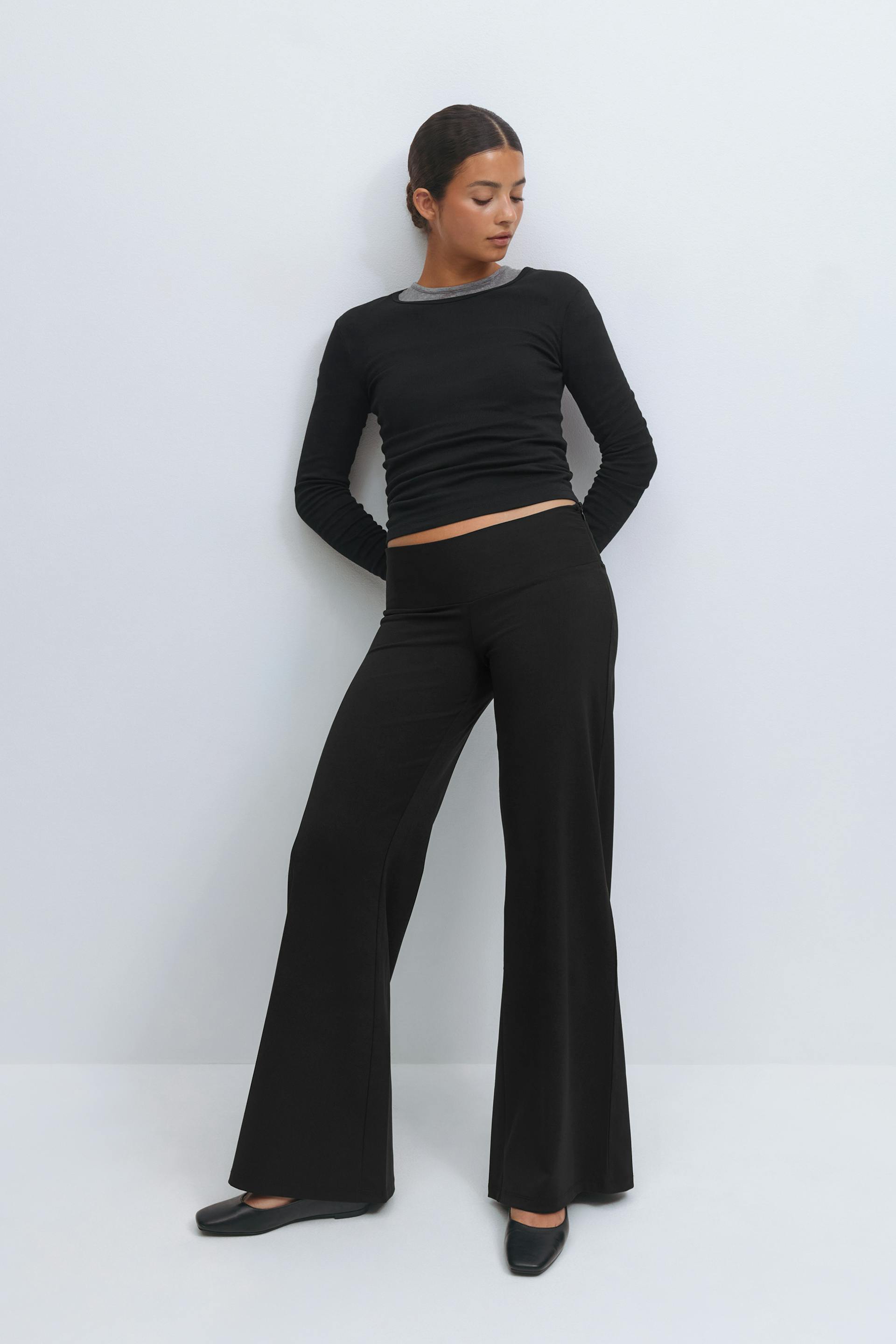 Black fitted long-sleeve top with a visible grey crew-neck underlayer, paired with high-waisted black wide-leg trousers and black square-toe ballet flats.