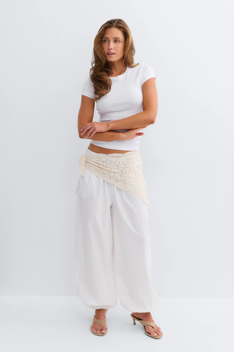 Witte top met korte mouwen, losse witte broek met wijde pijpen en elastiek in de zomen, crèmekleurige gehaakte taillesjaal vastgemaakt op de heup, beige teenslippers met hak.