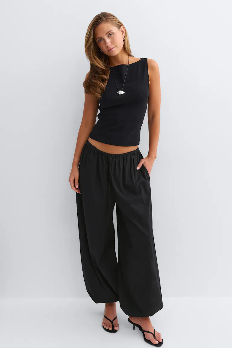 Aansluitende zwarte mouwloze top, zwarte broek met elastische taille, zwarte sandalen met hak en bandjes, lange hangerketting en kleine hoepeloorbellen.