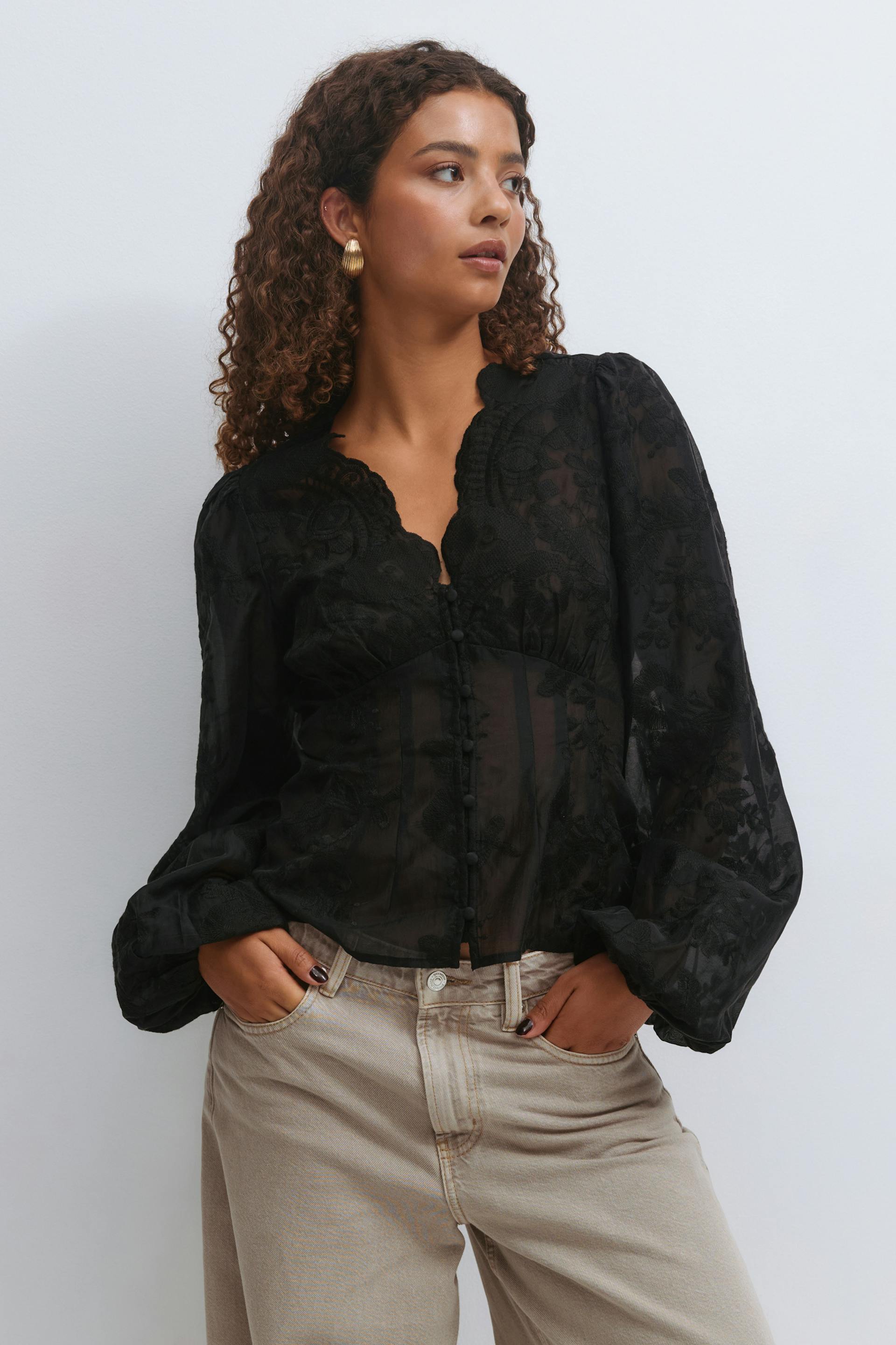 Svart, skjør, langermet bluse med broderi, takket V-utringning og stoffkledde knapper foran, kombinert med høytaljede, lysebeige jeans med avslappet passform.
