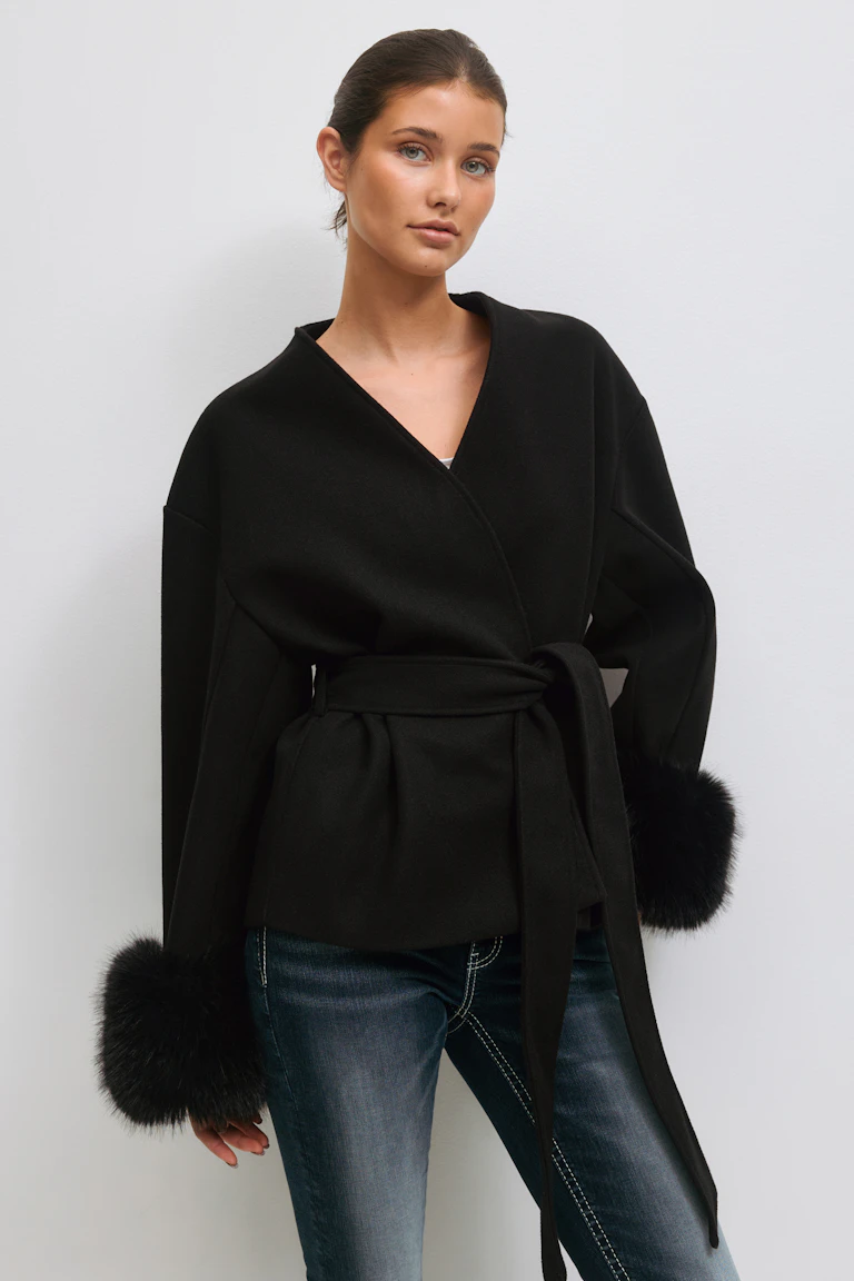 Schwarze Wickeljacke mit Gürtel, V-Ausschnitt und voluminösen Ärmeln mit schwarzen Pelzbündchen, die über ein weißes Oberteil gezogen wird; kombiniert mit verblichenen, schmal geschnittenen Bluejeans mit weißen Kontrastnähten.