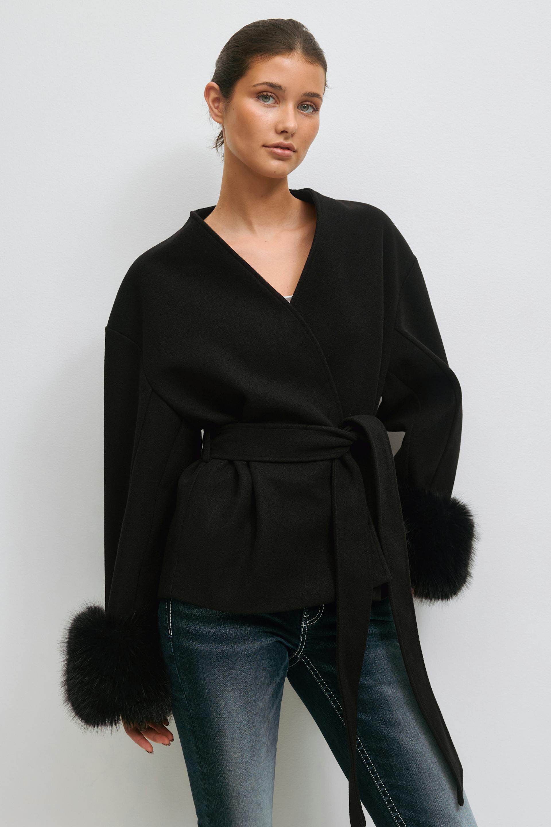 Black belted wrap jacket with V-neck and voluminous sleeves finished with black fur cuffs, layered over a white top; paired with faded slim-fit blue jeans with contrast white stitching.
