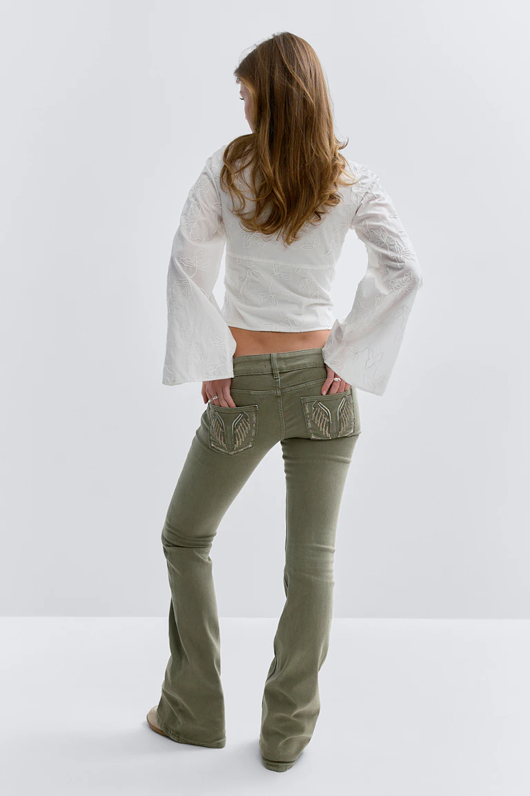 Witte kortgeknipte blouse met geborduurde bloemmotieven en wijde klokmouwen; olijfgroene uitlopende jeans met lage taille en versierde achterzakken.