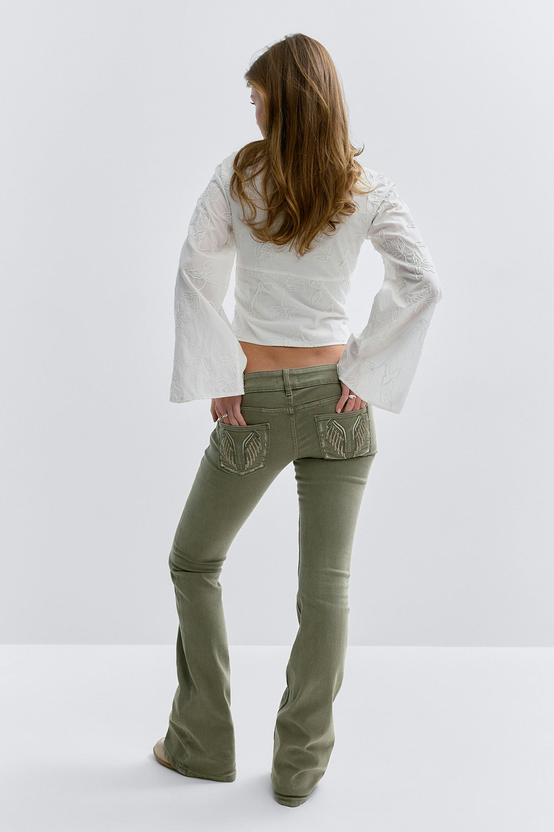 Weiße Kurzbluse mit Blumenstickerei und weiten Glockenärmeln; olivgrüne Low-Rise-Flared-Jeans mit verzierten Gesäßtaschen.