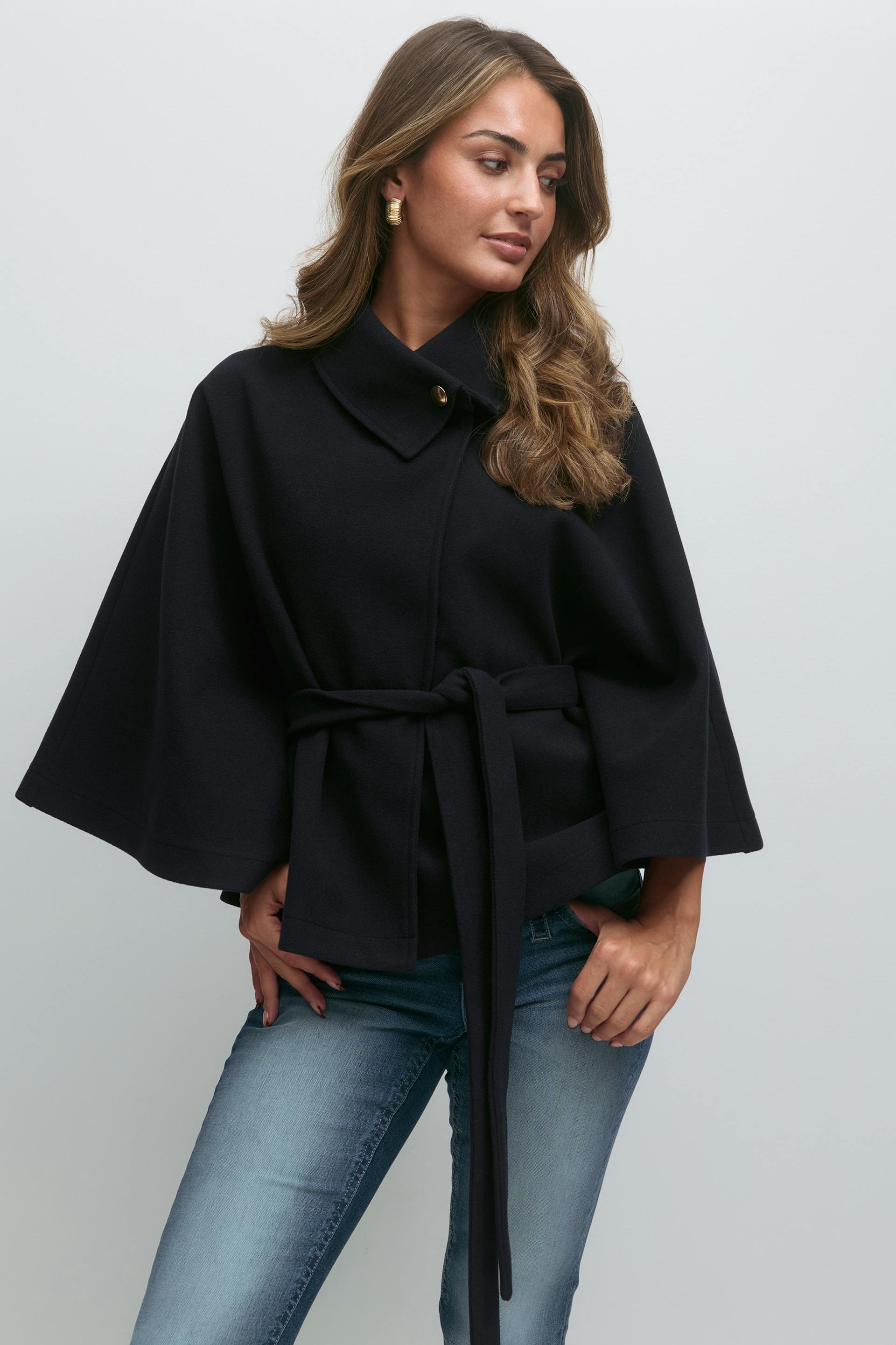 Navy belted cape coat with wide sleeves and a high collar fastened by a single gold button, paired with light-wash skinny jeans and gold hoop earrings.