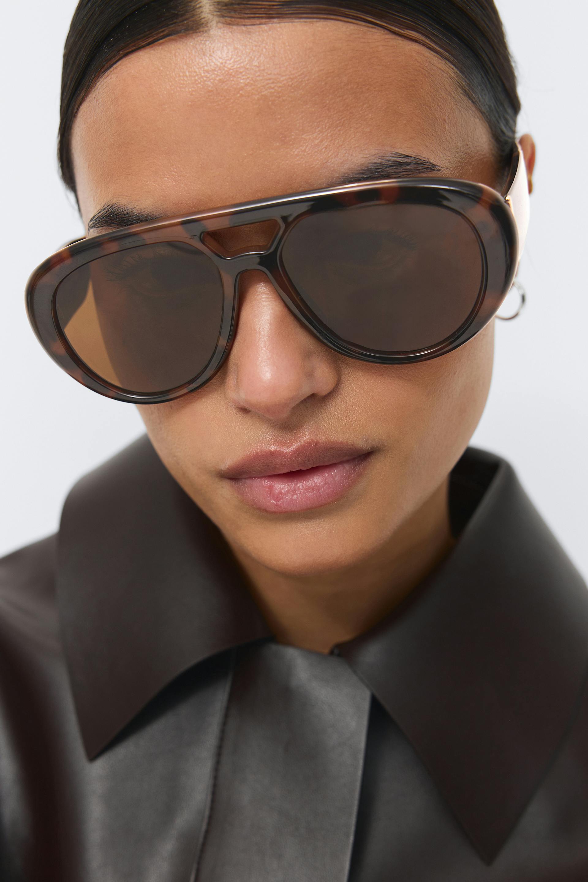 Übergroße Schildpatt-Sonnenbrille mit braunen Gläsern, kleiner silberner Ohrring und dunkelbraunes Lederhemd mit breitem Spitzkragen.