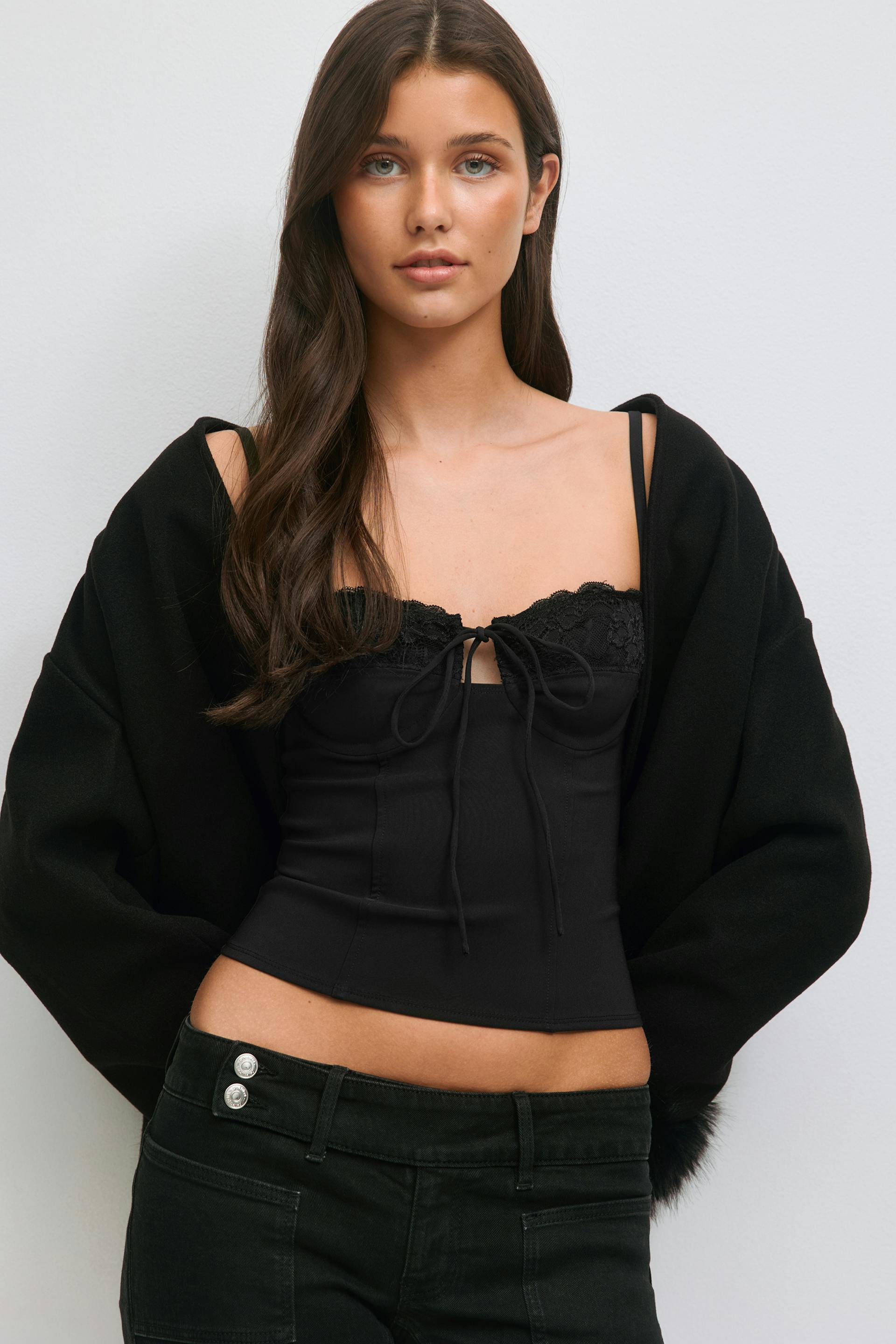 Black fitted corset-style camisole with lace-trimmed neckline, front tie and thin straps; oversized black jacket worn off the shoulders; black low-rise jeans with double silver waist buttons.