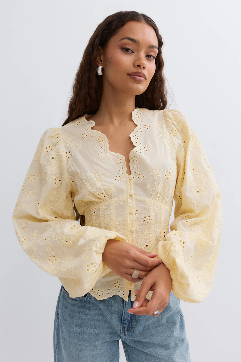Hellgelbe, bestickte Ösenbluse mit überschnittenem V-Ausschnitt, Knopfleiste und voluminösen Puffärmeln, kombiniert mit hellblauen Jeans.