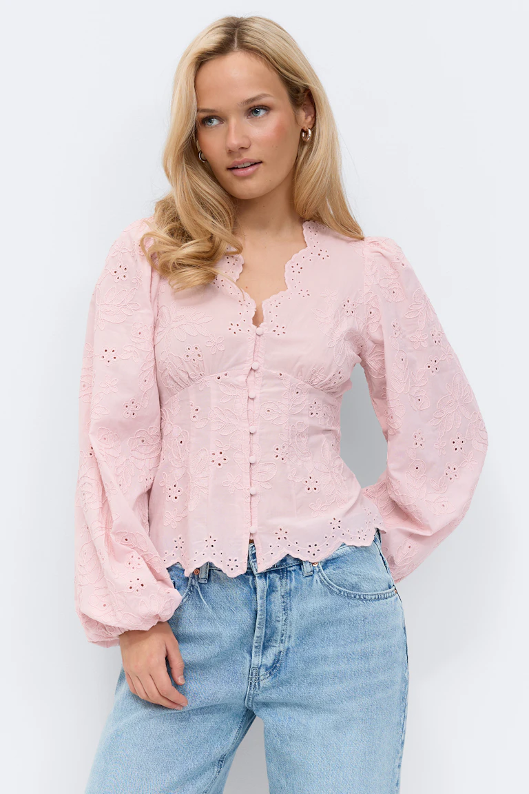 Lichtroze blouse met eyeletmotief en geschulpte V-hals, stoffen knopen aan de voorkant, lange pofmouwen en geschulpte zoom, gecombineerd met een lichtgekleurde blauwe jeans.