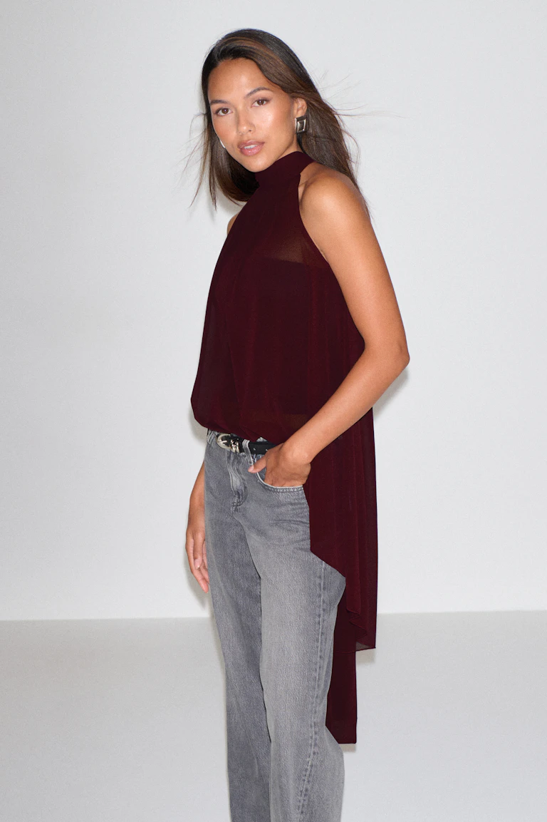 Ermeløs, skjør bluse i burgunder med høy hals og lang asymmetrisk drapering i ryggen, sammen med falmede, grå jeans med rette ben, et svart belte med sølvspenne og geometriske sølvøreringer.