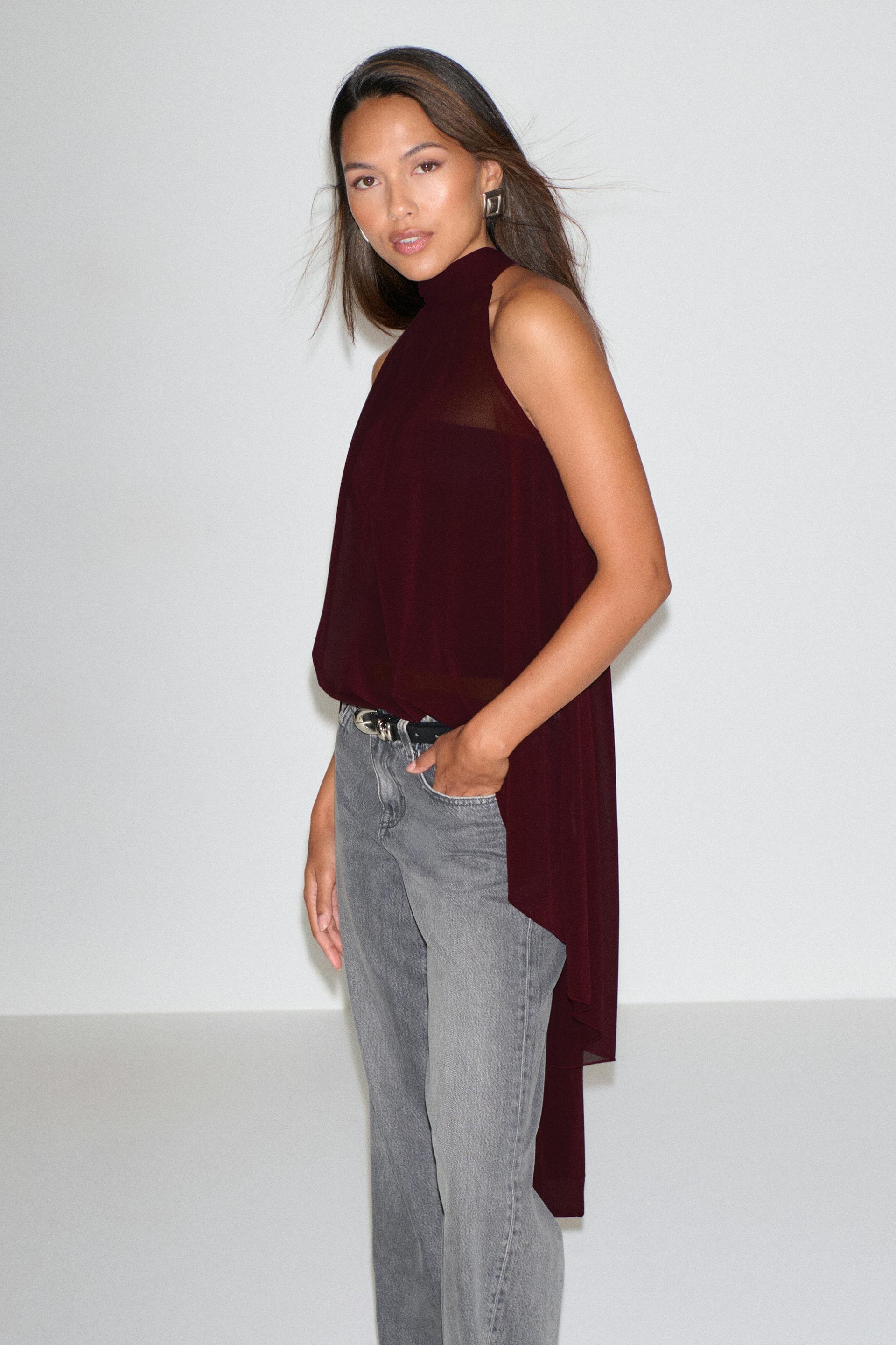 Läpikuultava, hihaton, korkeakaulainen viininpunainen pusero, jossa on pitkä, epäsymmetrinen selkäreunus, yhdistettynä haalistuneisiin harmaisiin suoralahkeisiin farkkuihin, mustaan vyöhön, jossa on hopeinen solki, ja geometrisiin hopeisiin korvakoruihin.