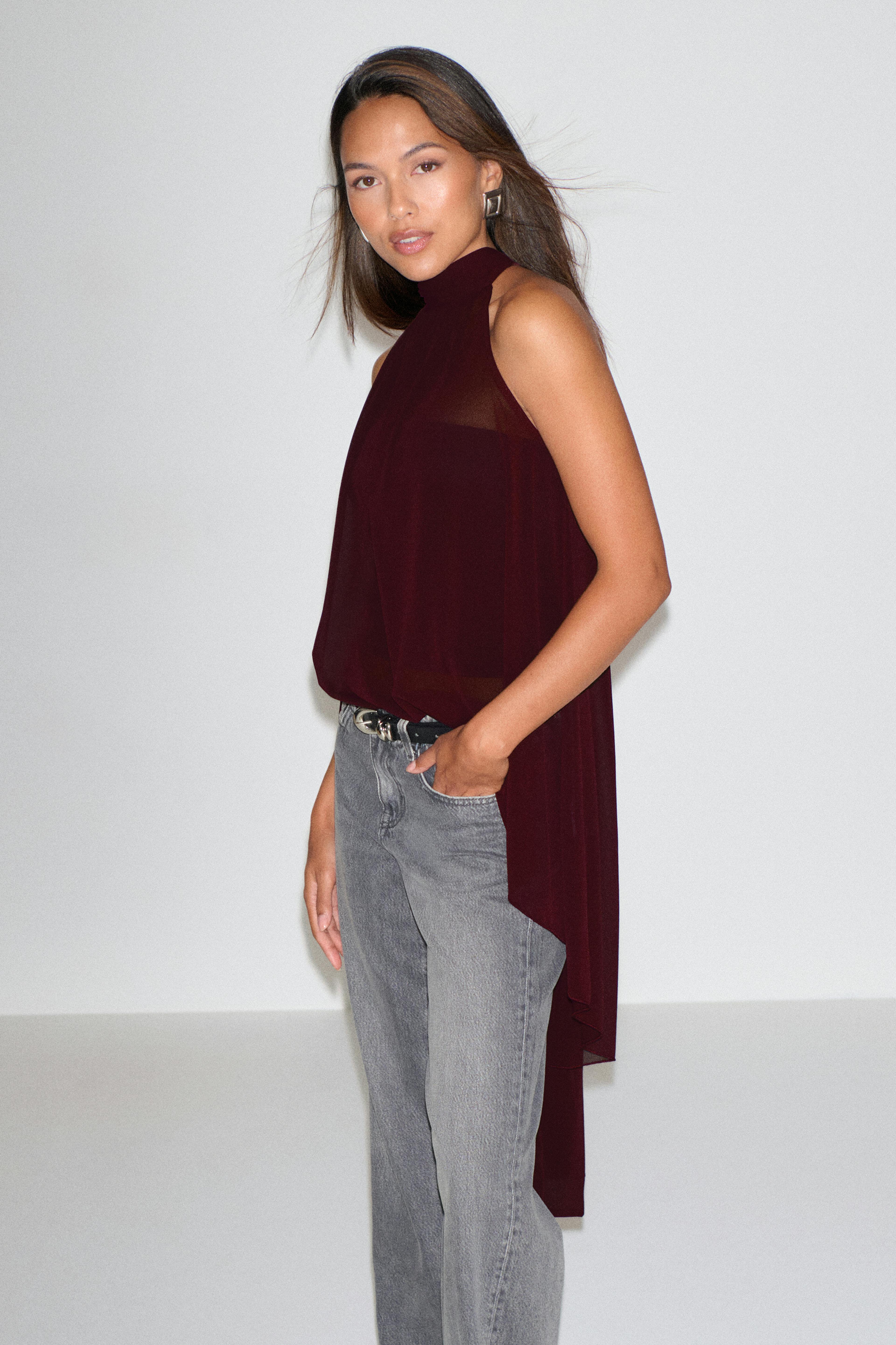 Donkerrode blouse zonder mouwen met hoge hals en een lange, asymmetrische drapering aan de achterkant, gecombineerd met een vaalgrijze jeans met rechte pijpen, een zwarte riem met zilveren gesp en geometrische zilveren oorbellen.