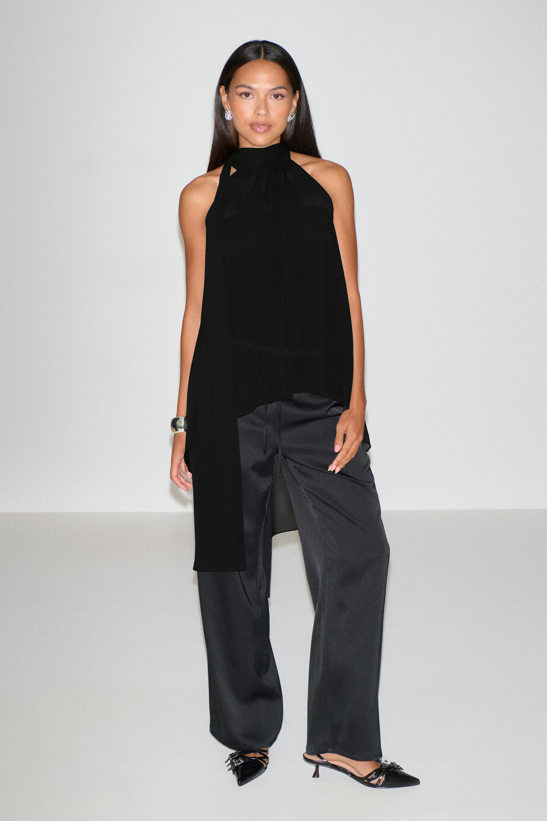 Sleeveless black halter-neck asymmetric tunic with a high neckline, paired with black wide-leg satin trousers and black pointed slingback kitten-heel mules; silver cuff bracelet and silver stud earrings.