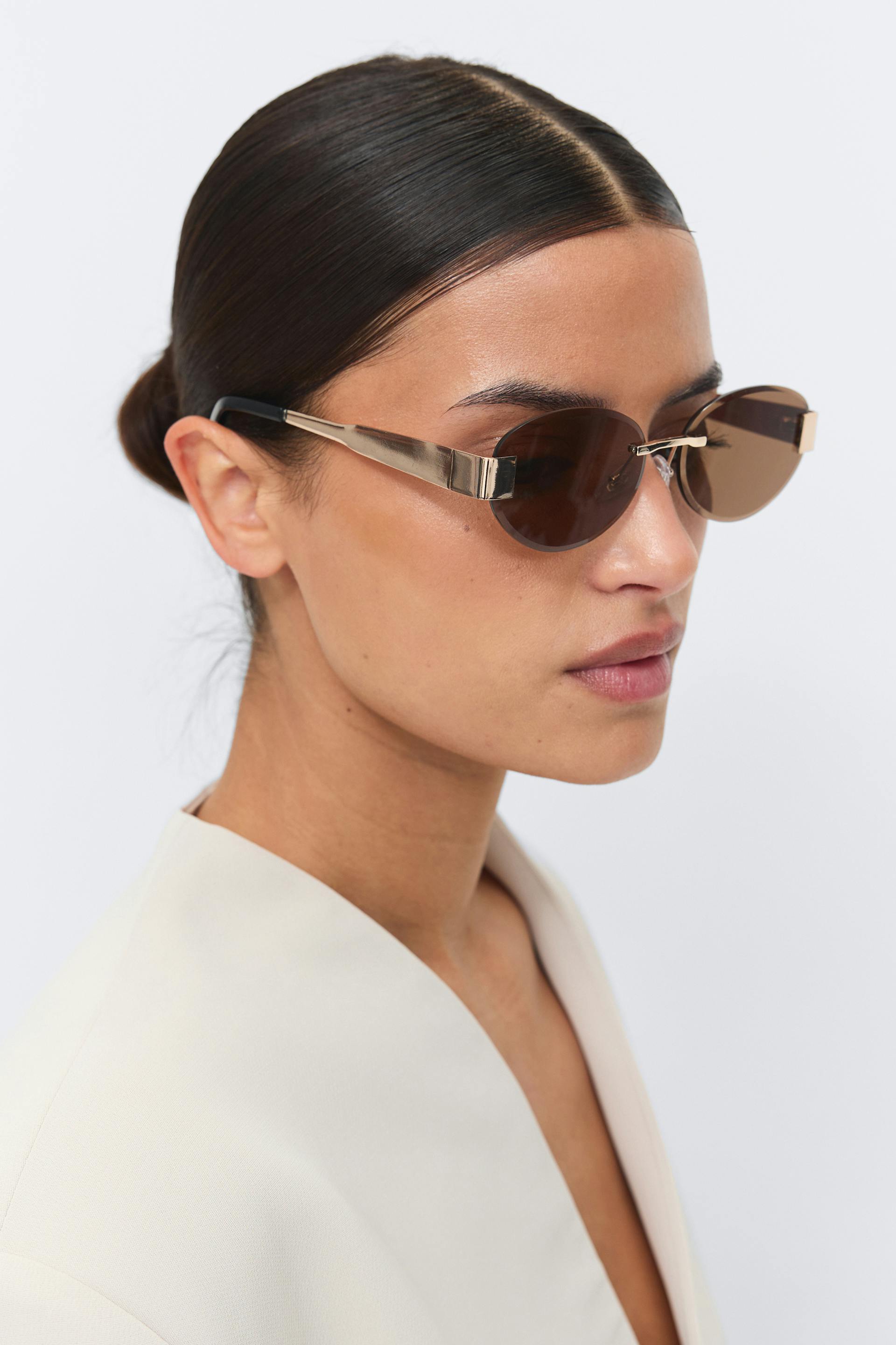 Off-white tailored blazer with a deep V neckline; slim oval brown-tinted sunglasses with a gold metal frame and broad gold temples finished with black tips.