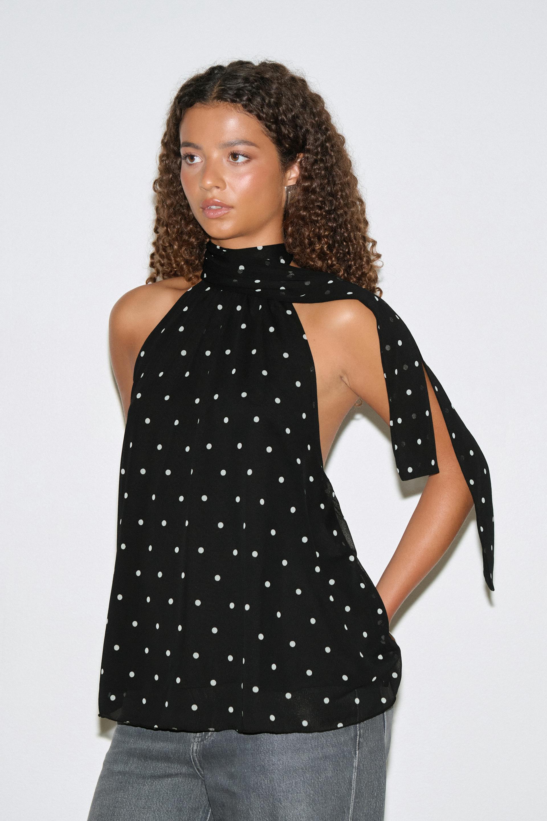 Musta hihaton halteripusero, jossa on valkoisia pilkkuja, korkeakauluksinen solmio ja olkapään huivi, yhdistettynä harmaisiin farkkuihin.