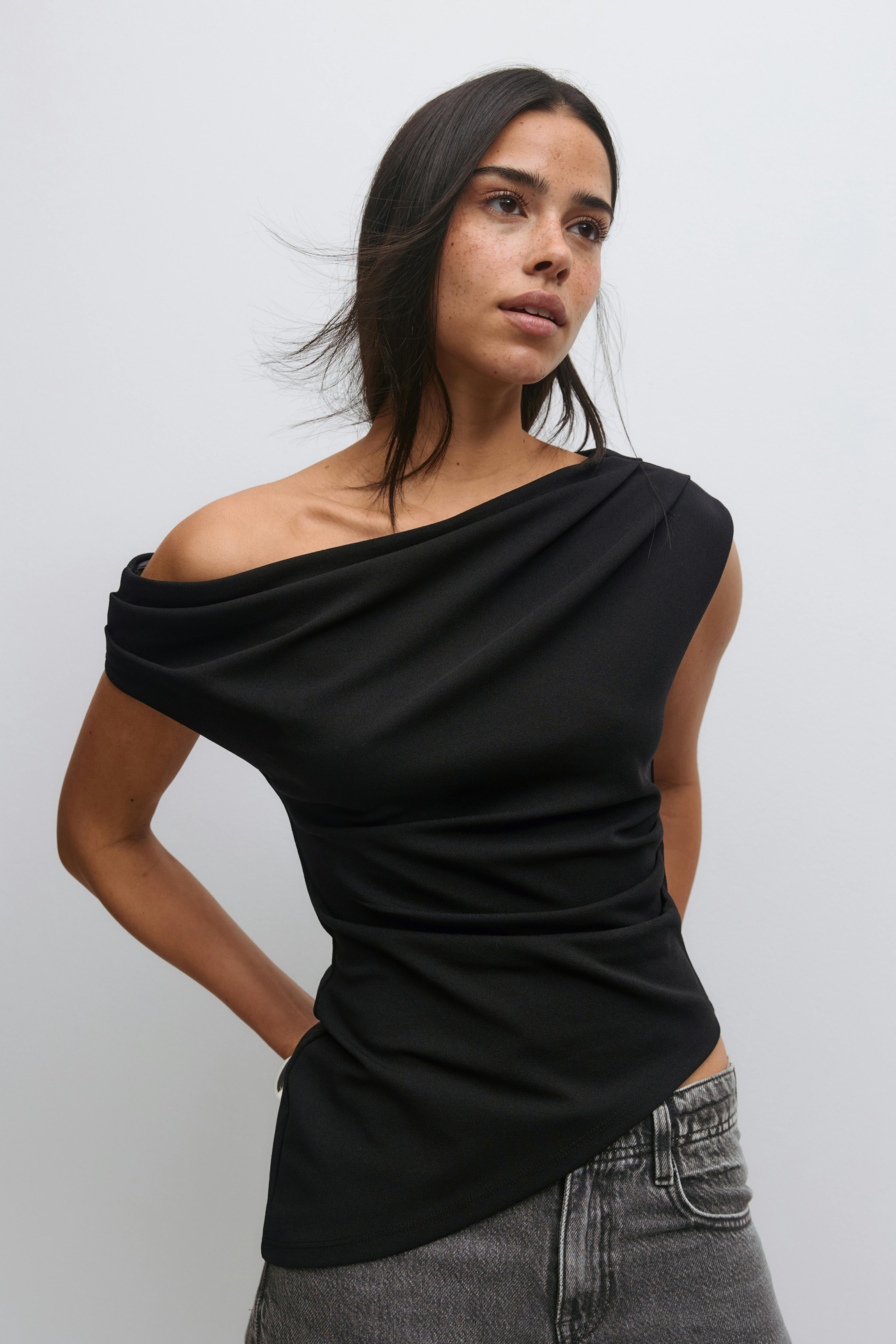 Zwarte asymmetrische gedrapeerde off-the-shoulder top met een enkele korte mouw, gecombineerd met een grijs jeans met hoge taille.