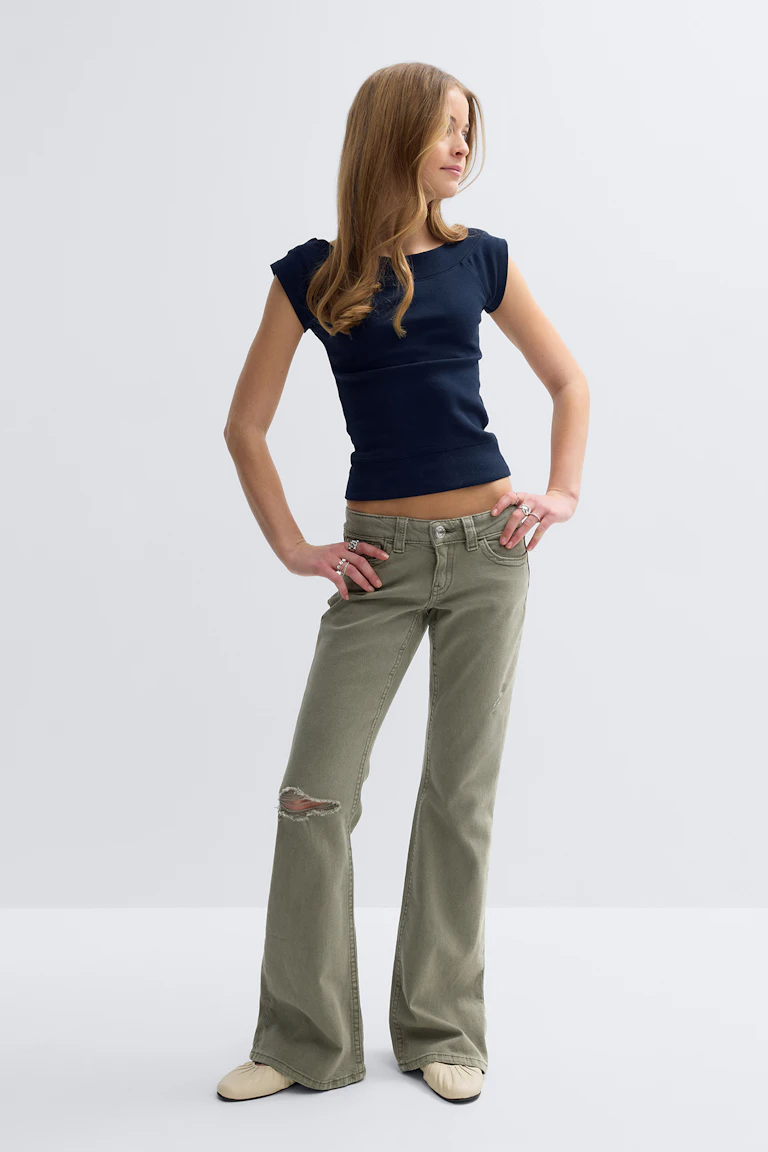 Navy fitted cap-sleeve top; olive-green low-rise flared jeans with a distressed rip at the left knee; beige slip-on flats; multiple rings on both hands.
