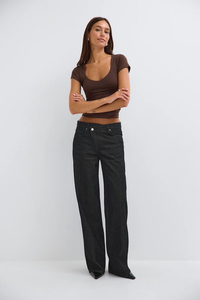 Elle porte un haut marron moulant à manches courtes et encolure dégagée, un jean noir à jambes larges et des escarpins noirs à bout pointu.