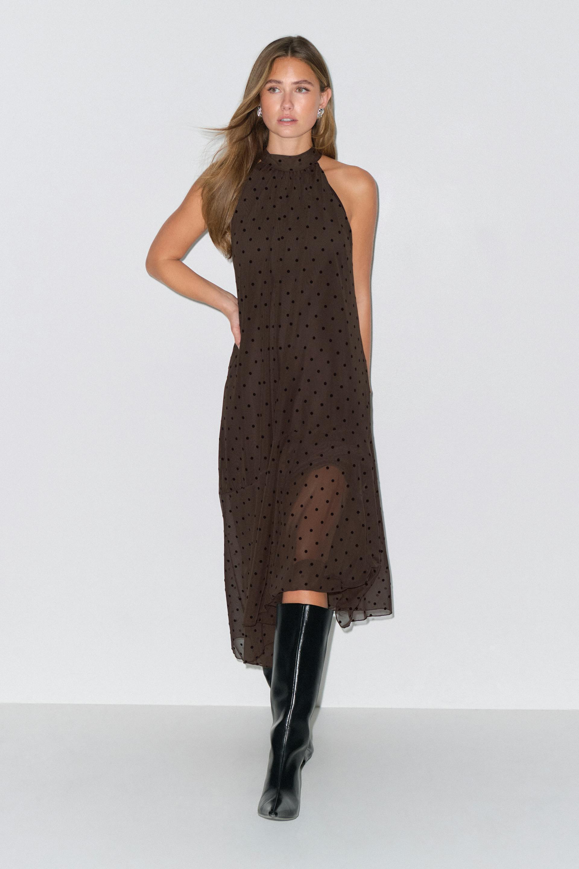 Sleeveless high‑neck brown midi dress with small black polka dots and sheer overlay, paired with knee‑high black leather boots.
