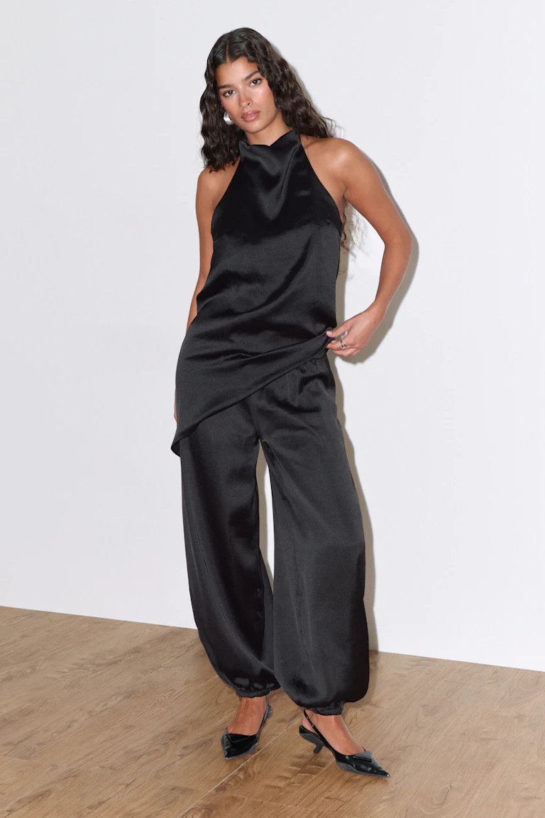 Schwarzes, drapiertes Oberteil aus Satin mit asymmetrischem Saum, kombiniert mit einer schwarzen, weit geschnittenen Jogginghose aus Satin mit elastischen Bündchen und schwarzen, spitzen Slingback-Absätzen.