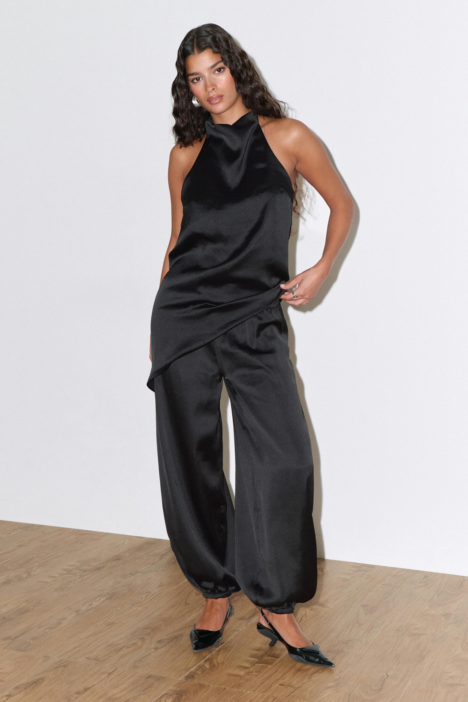Black satin halter-neck draped top with asymmetrical hem, paired with black satin wide-leg jogger trousers with elasticated cuffs and black pointed slingback heels.