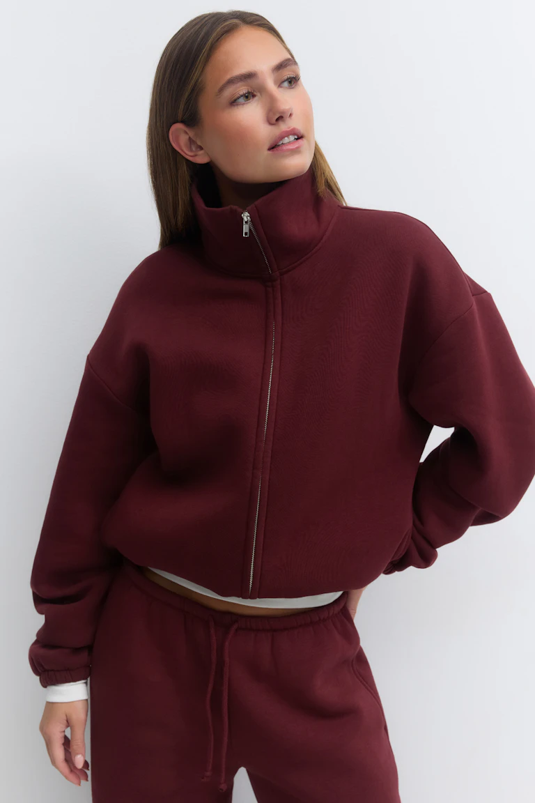 High-neck, full-zip cropped sweatshirt in deep burgundy with dropped shoulders and elastic cuffs, paired with matching burgundy drawstring sweatpants; white underlayer visible at cuffs and waist.