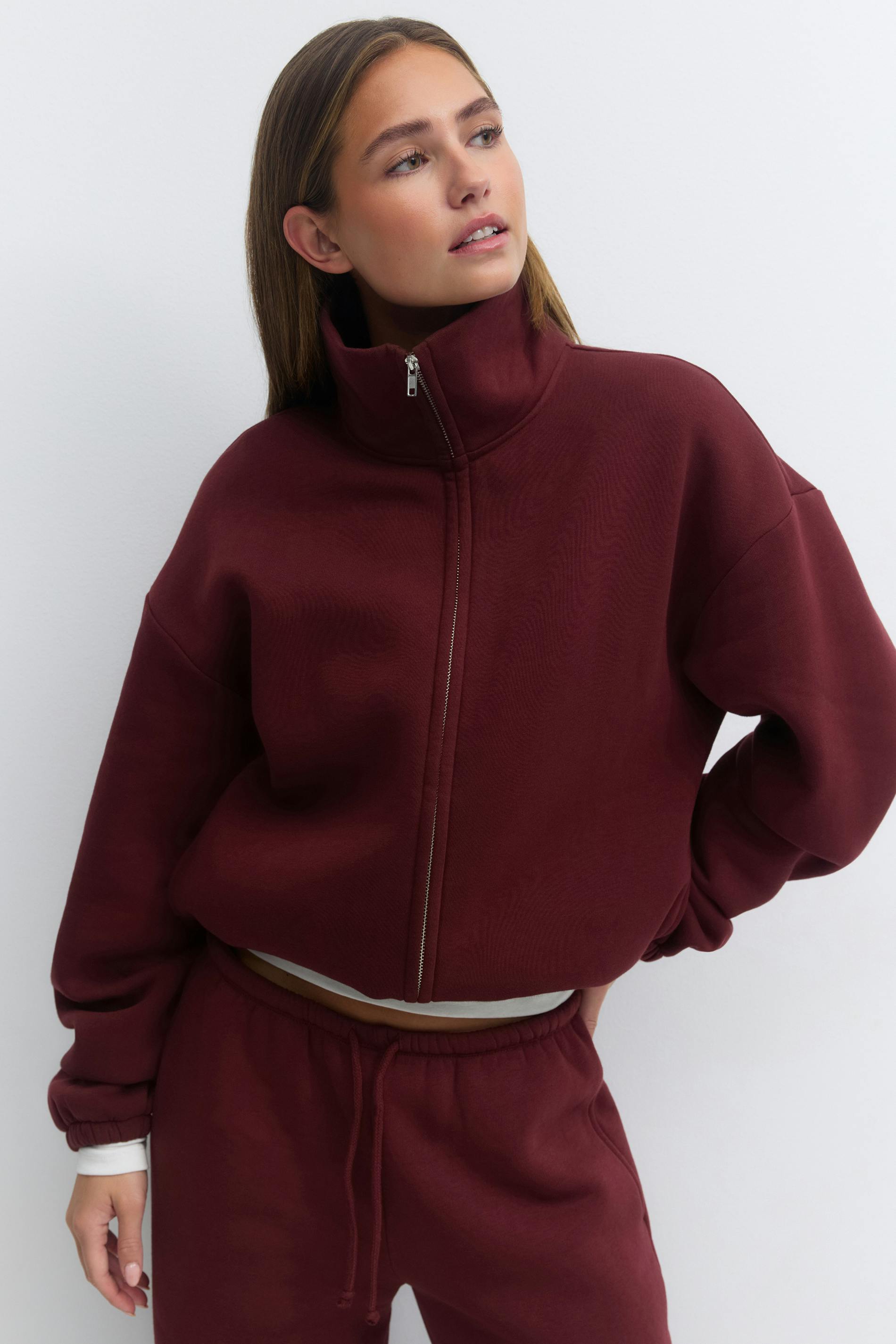 Korkeakaulainen, täysvetoketjullinen, syvän viininpunainen collegepaita, jossa on pudotetut olkapäät ja joustavat hihansuut, yhdistettynä yhteensopiviin viininpunaisiin vetoketjullisiin collegehousuihin; valkoinen aluskerros näkyy hihansuissa ja vyötäröllä.