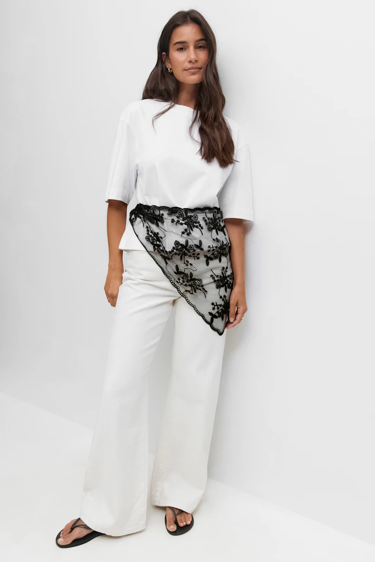 Witte boxy top met korte mouwen gecombineerd met een witte broek met wijde pijpen. Gecompleteerd met een zwart geborduurde kanten asymmetrische taillesjaal en zwarte teenslippers.