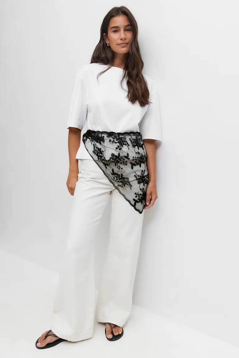 Witte boxy top met korte mouwen gecombineerd met een witte broek met wijde pijpen. Gecompleteerd met een zwart geborduurde kanten asymmetrische taillesjaal en zwarte teenslippers.