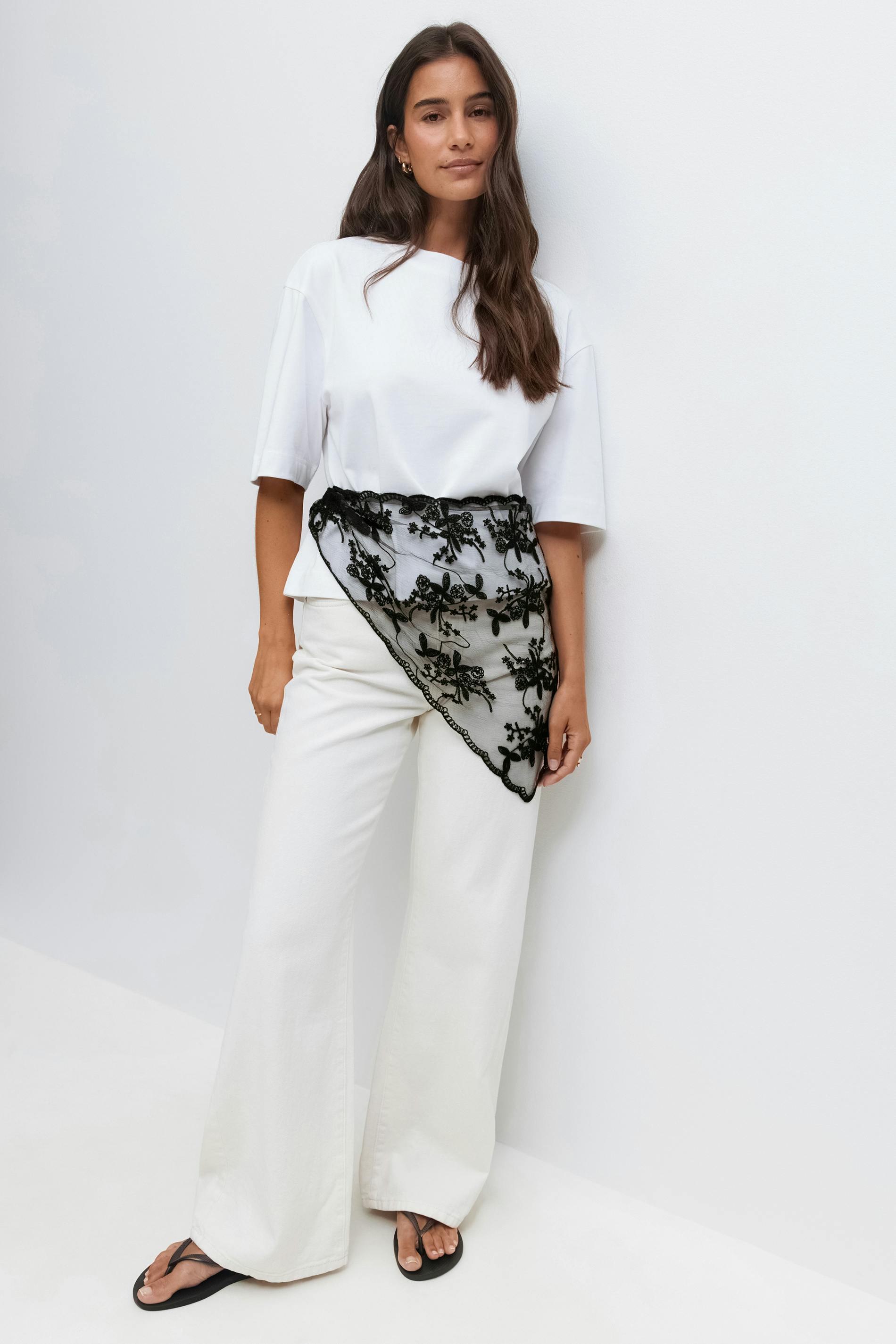 White short-sleeve boxy top paired with white wide-leg trousers, accessorised with a black embroidered lace asymmetrical waist scarf and black thong sandals.