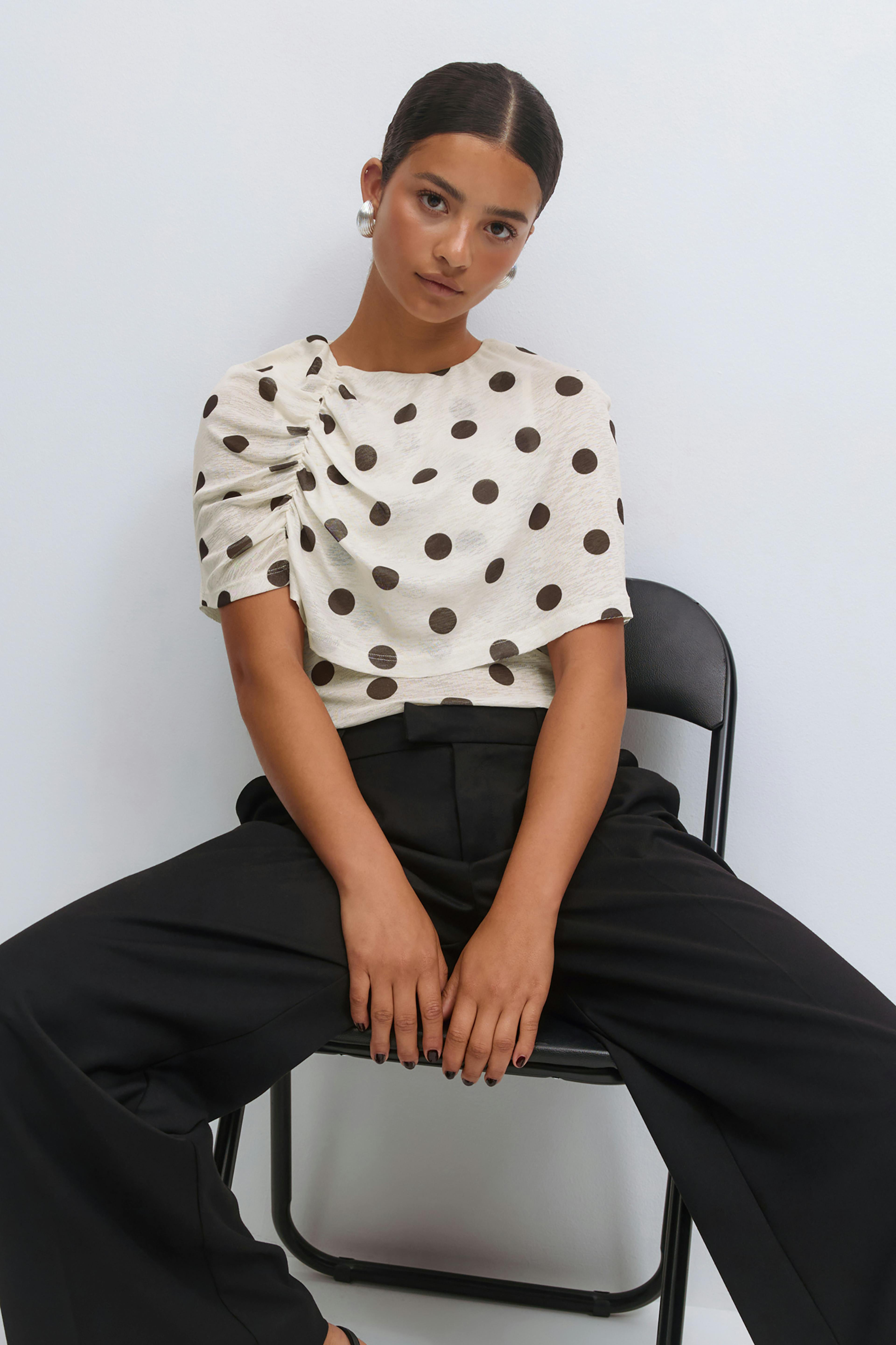 Cream short-sleeve blouse with large black polka dots and an asymmetric ruched shoulder detail, paired with high-waisted black wide-leg trousers and silver hoop earrings.