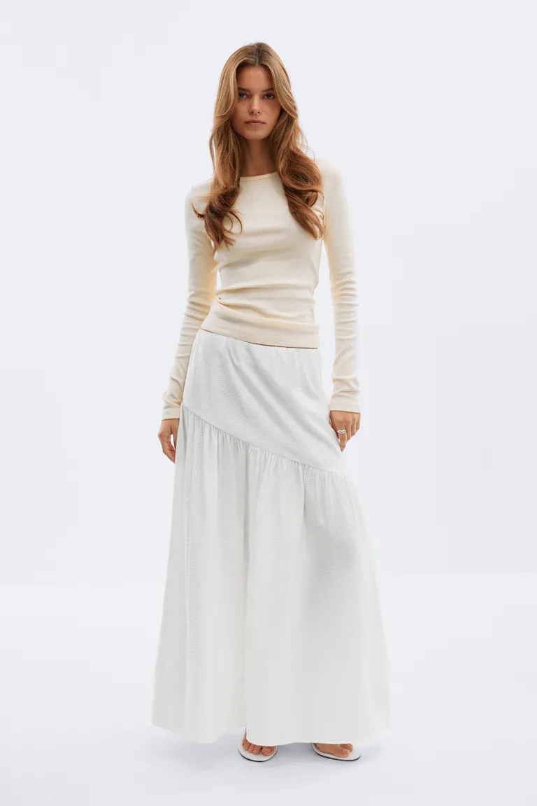 Fitted cream long-sleeve crewneck top paired with an off-white asymmetrical, tiered maxi skirt and white open-toe sandals.