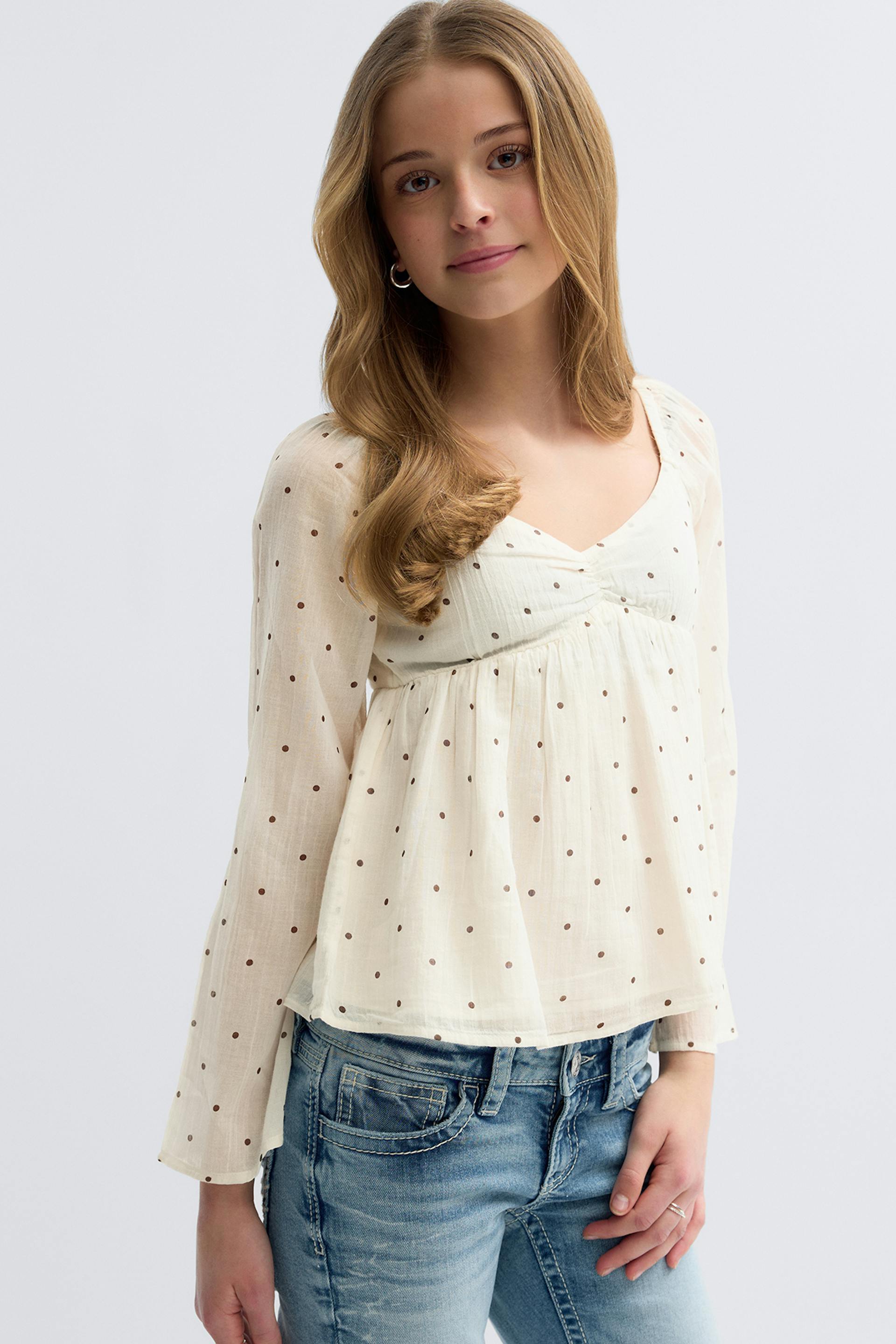Ivory long-sleeve V-neck semi-sheer blouse with small brown polka dots, an empire seam and gathered peplum hem, paired with light-wash blue jeans.