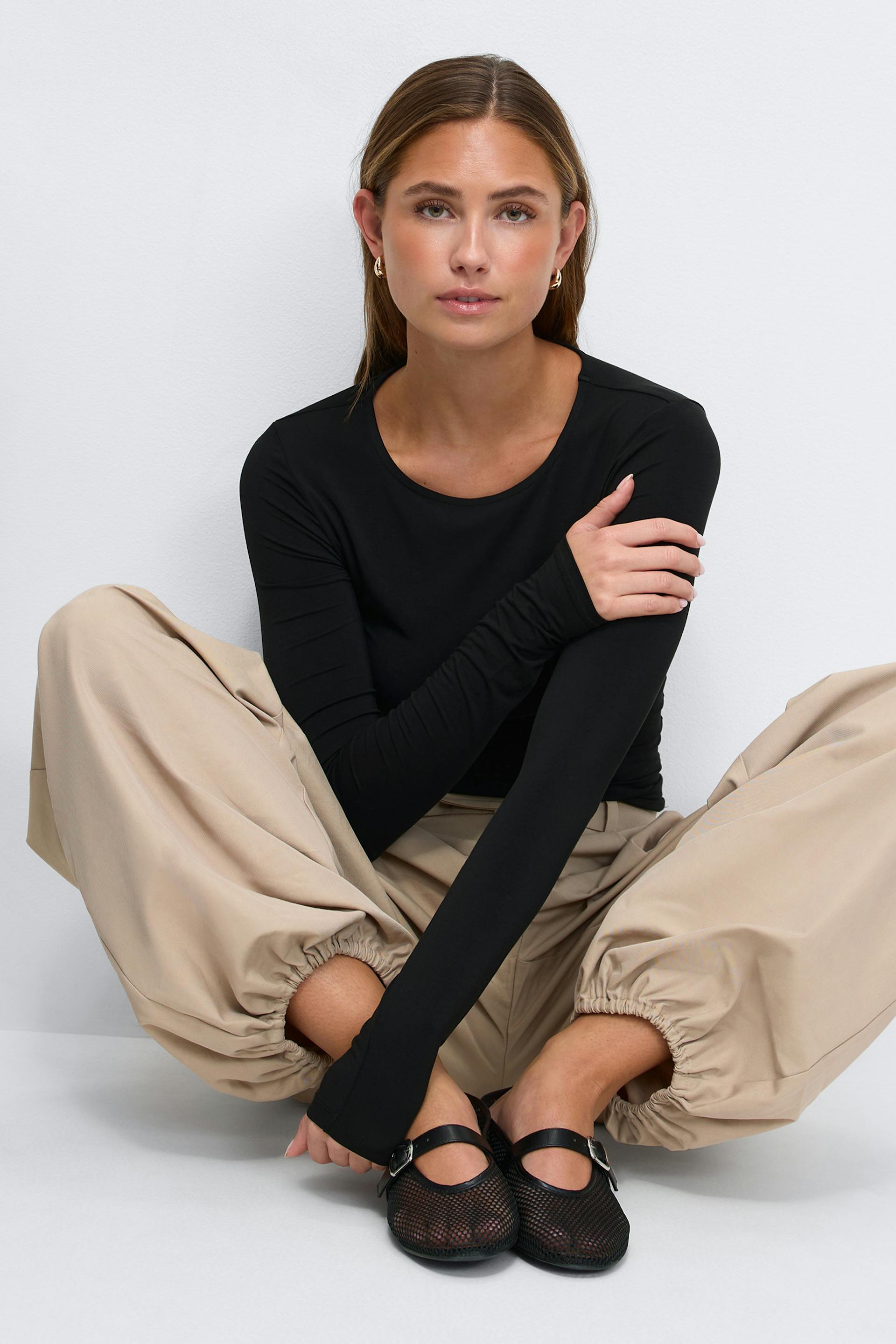 Svart långärmad topp med rund halsringning, beige byxor med vida ben och elastiska muddar, svarta Mary Jane-flats i mesh med spänne och små örhängen i guld.