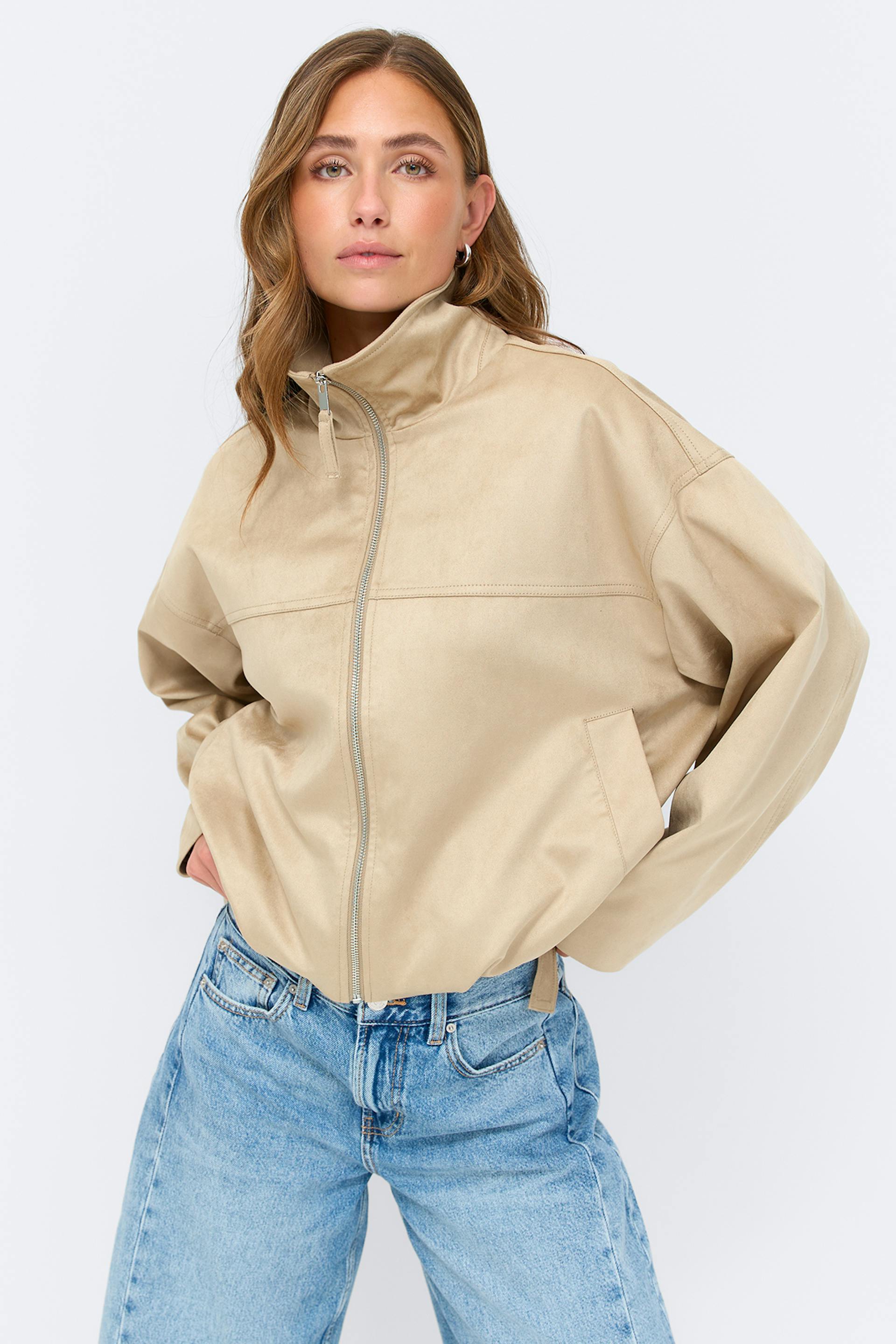 Beige vetoketjullinen takki, jossa on korkea kaulus, pitkät hihat ja sivutaskut, yhdistettynä vaaleaksi pestyihin, korkeavyötäröisiin, suoralinjaisiin farkkuihin.