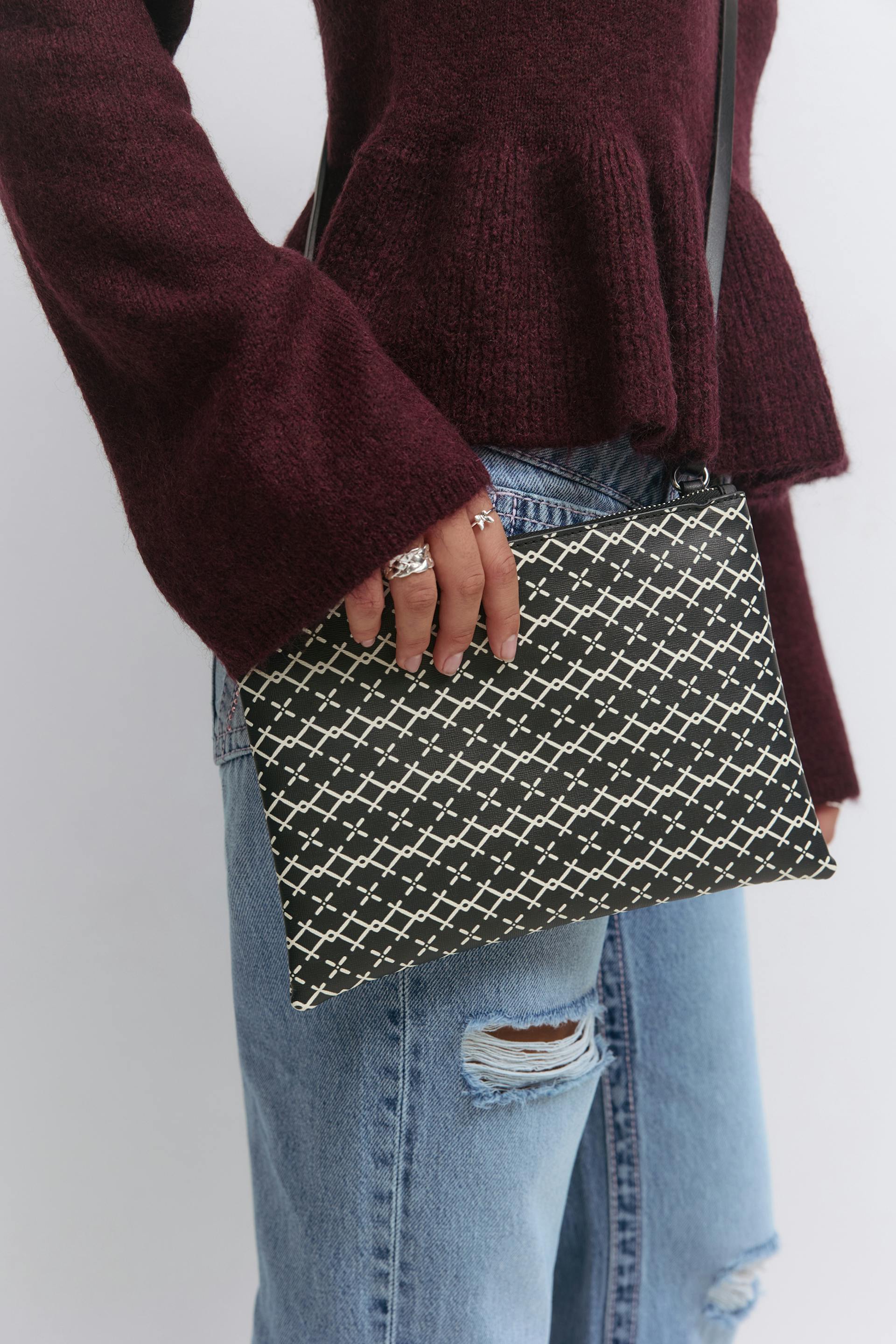 Burgundy knit sweater, light-wash distressed jeans with knee rips, black crossbody bag with white lattice pattern, and silver rings.