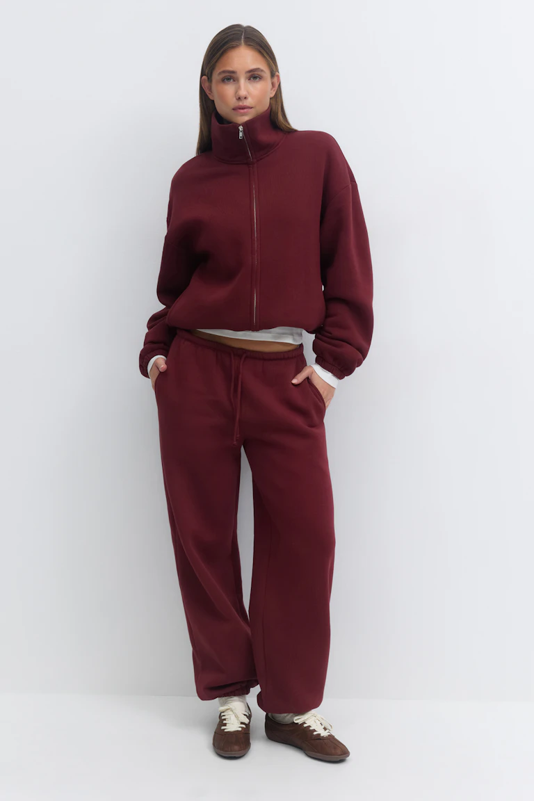 Woman wearing a burgundy funnel‑neck zip jacket paired with matching drawstring wide‑leg sweatpants, a white T‑shirt peeking at the hem, and brown lace‑up sneakers.