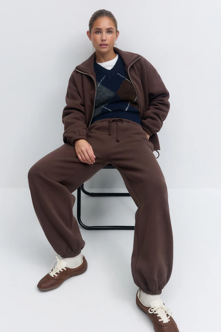 Veste zippée marron à poignets élastiques, pantalon de jogging marron à cordon de serrage assorti, pull en argyle marine à col en V superposé à un T-shirt blanc à col ras du cou, chaussettes blanches et baskets marron à lacets.