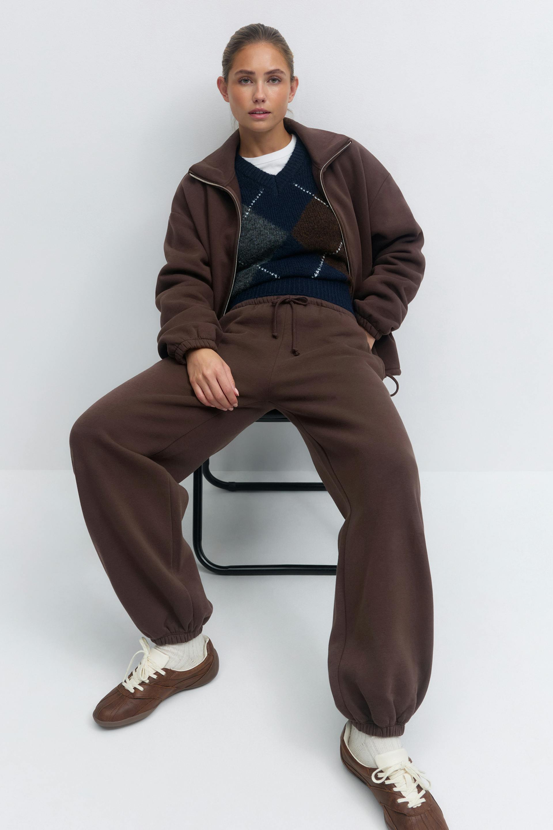 Braune Jacke mit Reißverschluss und elastischen Bündchen, dazu passende braune Jogginghose mit Kordelzug, marineblauer Pullover mit V-Ausschnitt über einem weißen T-Shirt mit Rundhalsausschnitt, weiße Socken und braune Schnürschuhe.