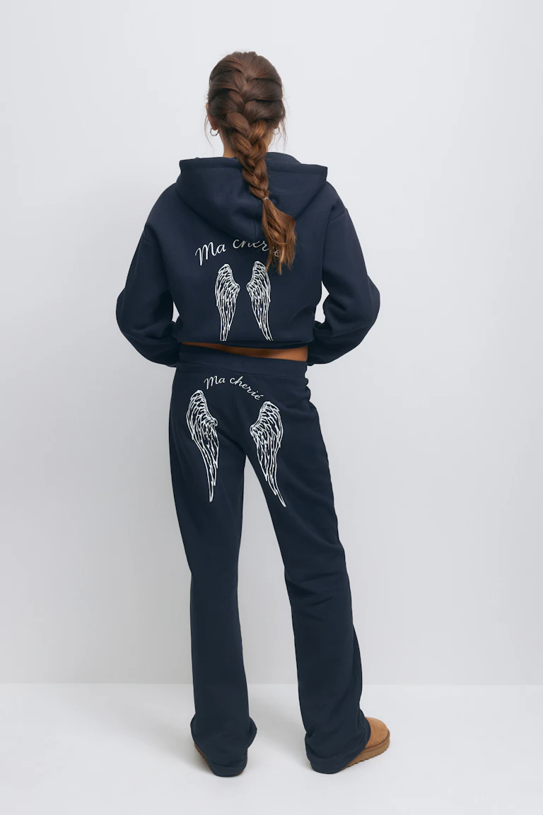 Marineblaues Kapuzensweatshirt mit weißer Engelsflügelgrafik und dem Schriftzug "Ma chérie" auf dem Rücken, kombiniert mit einer passenden marineblauen Jogginghose mit weißer Flügelgrafik und dem Schriftzug "Ma chérie" auf dem Gesäß sowie hellbraunen Slippern aus Wildleder.