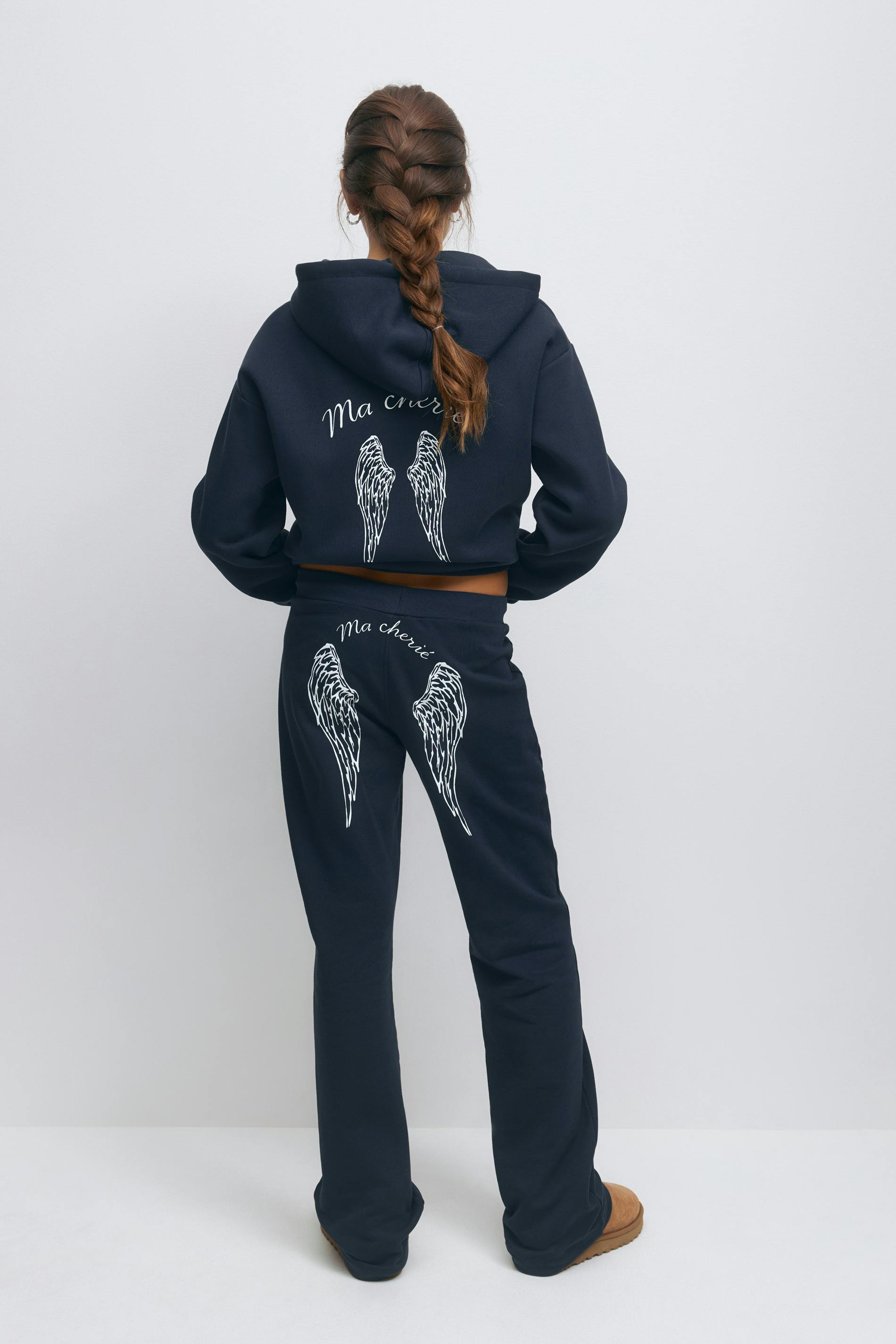 Navy cropped hooded sweatshirt with white angel‑wing graphic and "Ma chérie" script on the back, paired with matching navy sweatpants featuring white wing graphics and "Ma chérie" script on the seat, and tan slip-on suede slippers.