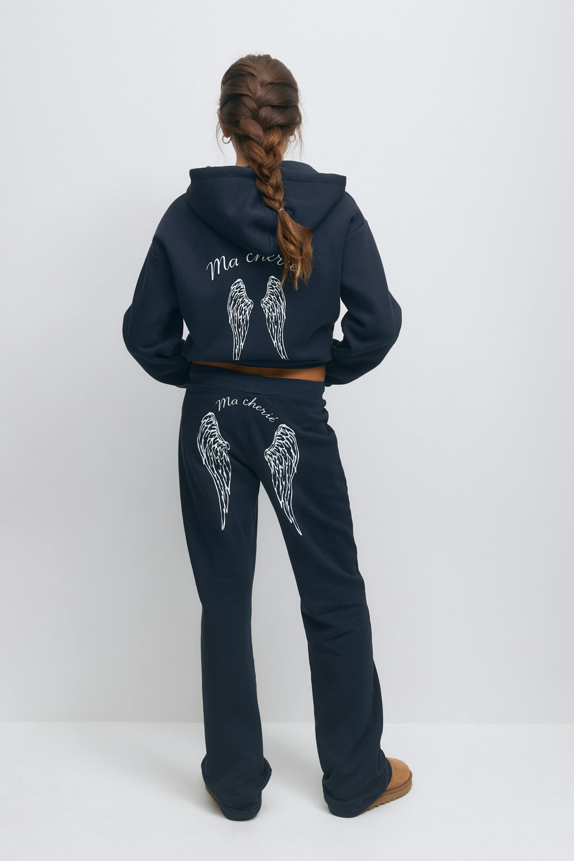 Navy cropped hooded sweatshirt with white angel‑wing graphic and "Ma chérie" script on the back, paired with matching navy sweatpants featuring white wing graphics and "Ma chérie" script on the seat, and tan slip-on suede slippers.